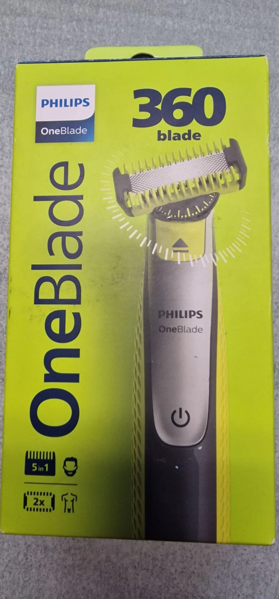 Opakowanie golarki Philips OneBlade w kolorze jasnozielonym. Na przodzie znajduje się duży pionowy napis 'OneBlade' oraz napis '360 blade'. Widoczne jest zdjęcie urządzenia z głowicą golącą i logo PHILIPS OneBlade na obudowie. W lewym dolnym rogu znajdują się ikony informujące o zawartości zestawu: '5 in 1' oraz '2x' przy symbolu ostrza.