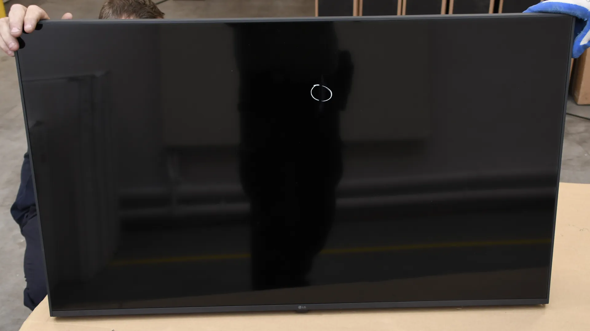 Zdjęcie przedstawiające czarny, błyszczący ekran telewizora lub monitora widziany od przodu. Na środku ekranu znajduje się mały, biały okrąg. W odbiciu ekranu widoczna jest sylwetka osoby. W górnym lewym rogu widoczna jest dłoń trzymająca krawędź urządzenia. Na dolnej ramce znajduje się małe logo LG.
