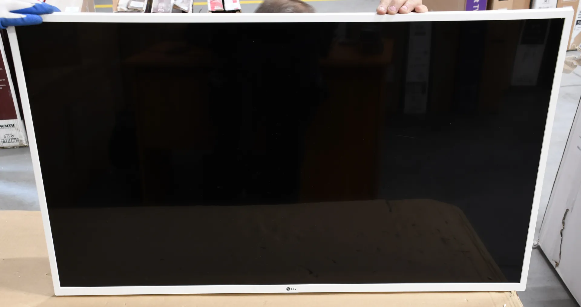 Zdjęcie przedstawiające czarny ekran monitora lub telewizora w białej ramce, trzymany przez dwie osoby. Na dolnej krawędzi ramki znajduje się małe logo LG. Ekran jest wyłączony i odbija otoczenie.