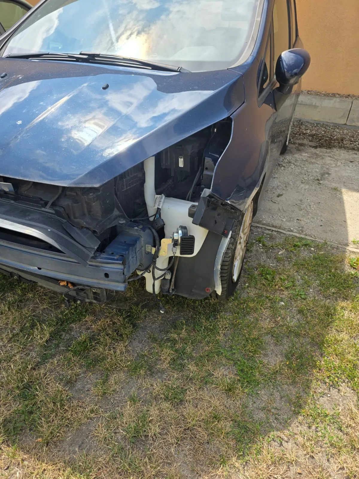 Zbliżenie na przód uszkodzonego, ciemnoniebieskiego samochodu. Brakuje przedniego reflektora po prawej stronie oraz części zderzaka, co odsłania wewnętrzne elementy konstrukcyjne pojazdu, w tym białe plastikowe obudowy i przewody. Samochód stoi na trawiasto-piaszczystym podłożu obok betonowego krawężnika.