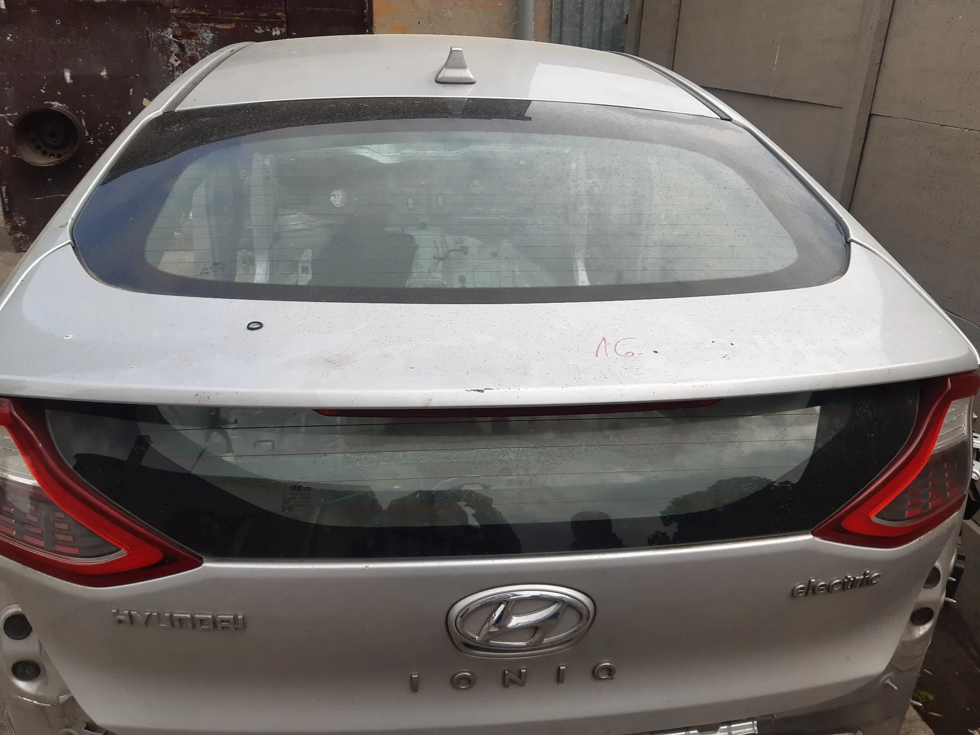 Tylny widok srebrnego samochodu marki Hyundai Ioniq Electric. Widoczna jest tylna szyba, klapa bagażnika z logo Hyundai oraz napisami 'HYUNDAI', 'IONIQ' i 'electric'. Na karoserii nad tylną szybą znajduje się czerwony napis '16'.