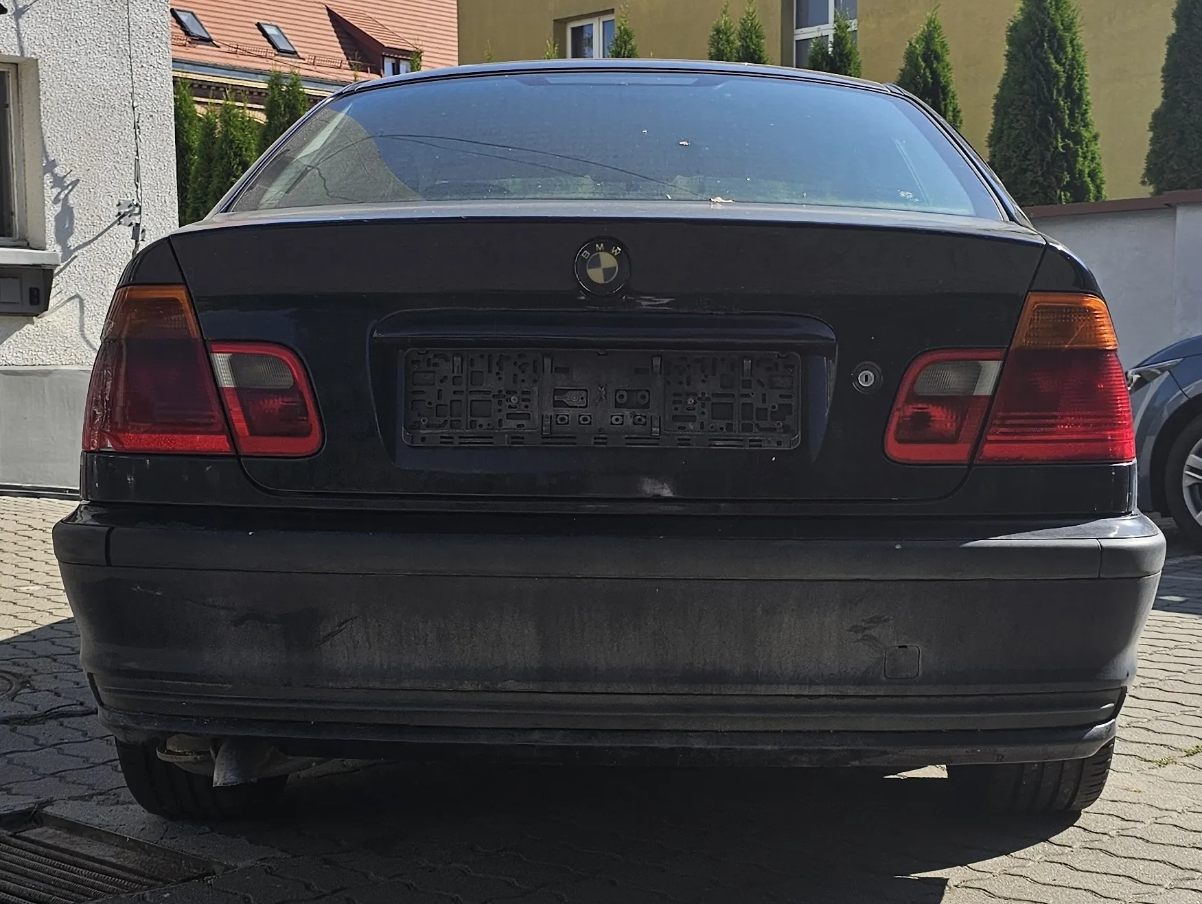 Tylny widok czarnego samochodu marki BMW. Pojazd nie posiada tablic rejestracyjnych, widoczne jest jedynie puste miejsce na ich montaż. Samochód jest zakurzony, a na zderzaku widoczne są zabrudzenia. W tle widać fragmenty budynków oraz zielone drzewa iglaste.