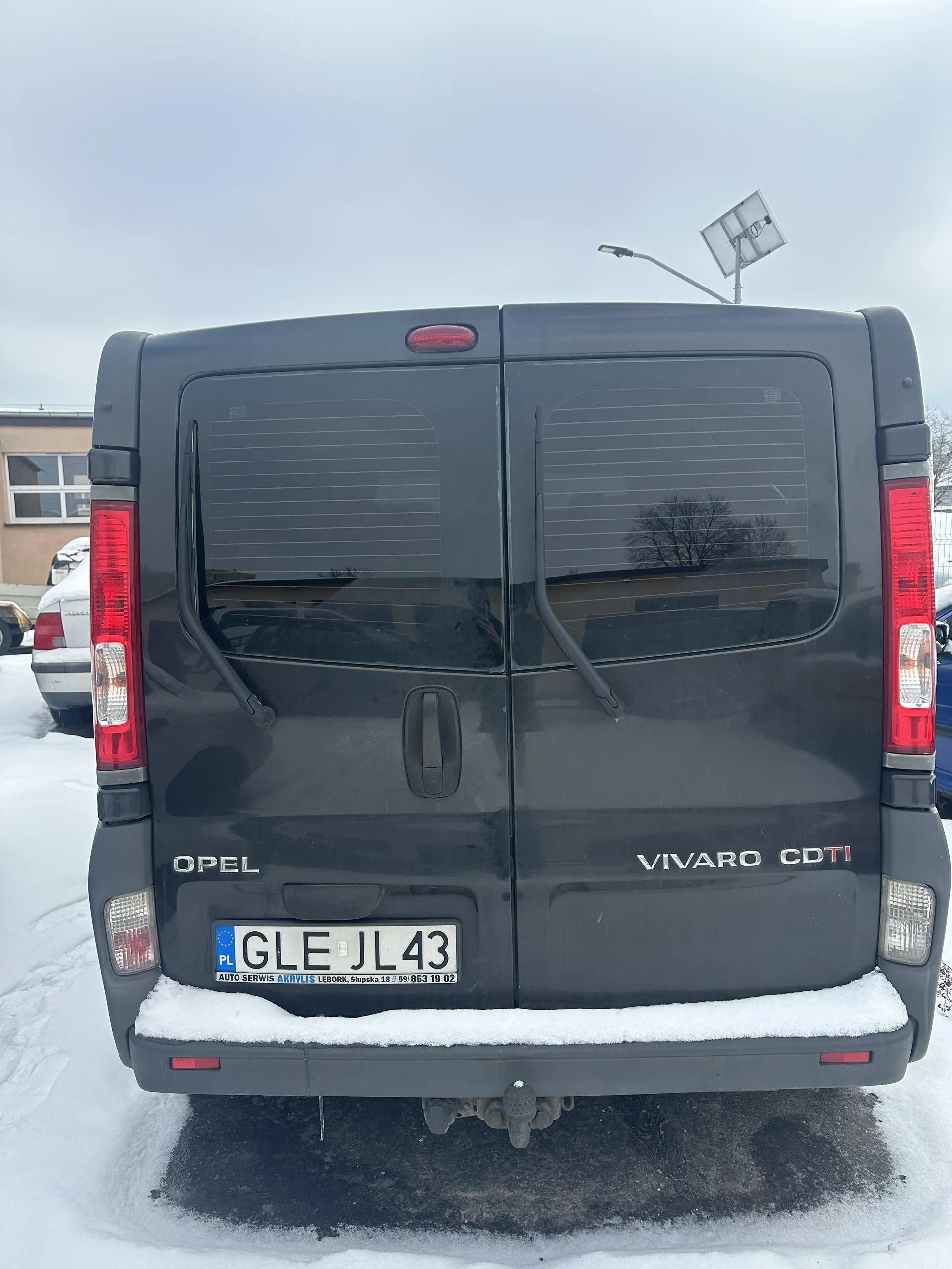 Tył ciemnoszarego samochodu dostawczego Opel Vivaro CDTI zaparkowanego na śniegu. Widoczne są czerwone światła tylna, tablica rejestracyjna GLE JL 43 z niebieskim pasem PL po lewej stronie oraz napisem AUTO SERWIS AKRVIS LEBORK, Słupska 18 59/663 19 02 pod nią. Na lewej stronie tyłu napis OPEL, na prawej VIVARO CDTI. Tylne drzwi z przyciemnionymi szybami i poziomymi liniami. W tle budynek z oknami, częściowo zakryty śniegiem samochód, latarnia uliczna i panel słoneczny. Niebo jest pochmurne.