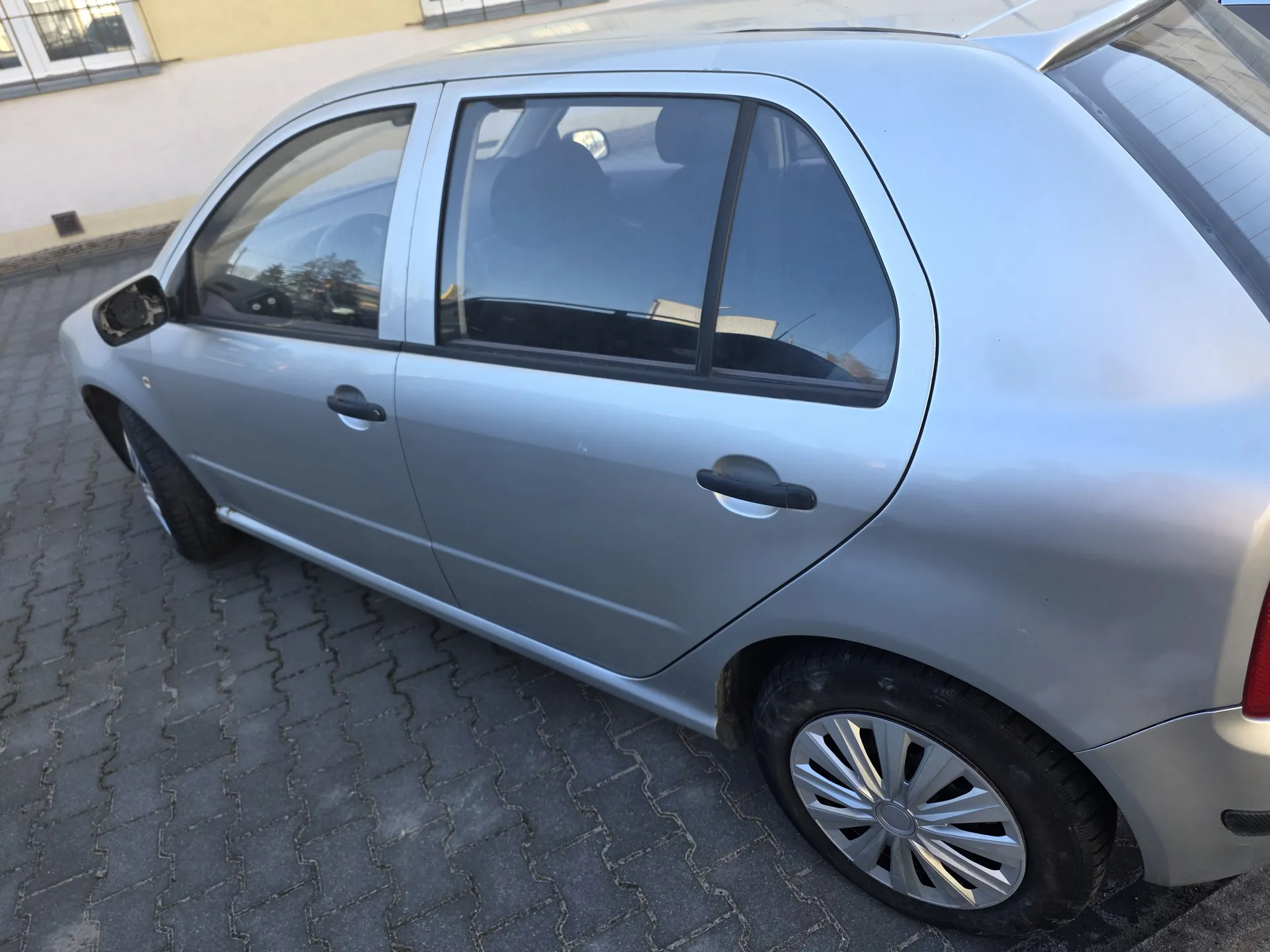 Srebrny samochód osobowy typu hatchback zaparkowany na brukowanej powierzchni. Widoczne są boczne drzwi, okna, lusterko boczne oraz felgi. W tle budynek z żółtą ścianą i oknami. Samochód stoi na płaskim terenie, a horyzont jest poziomy.