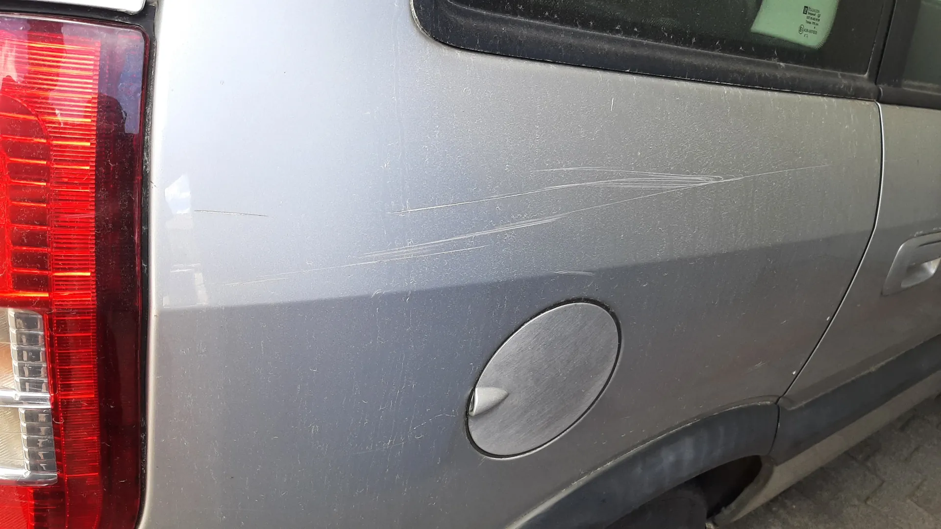 Zbliżenie na tylną część srebrnego samochodu. Widoczny czerwony reflektor z lewej strony, metalowa pokrywa zbiornika paliwa oraz wyraźne, długie zadrapania na karoserii. Na tle fragment asfaltu i okno z zielonym naklejką.