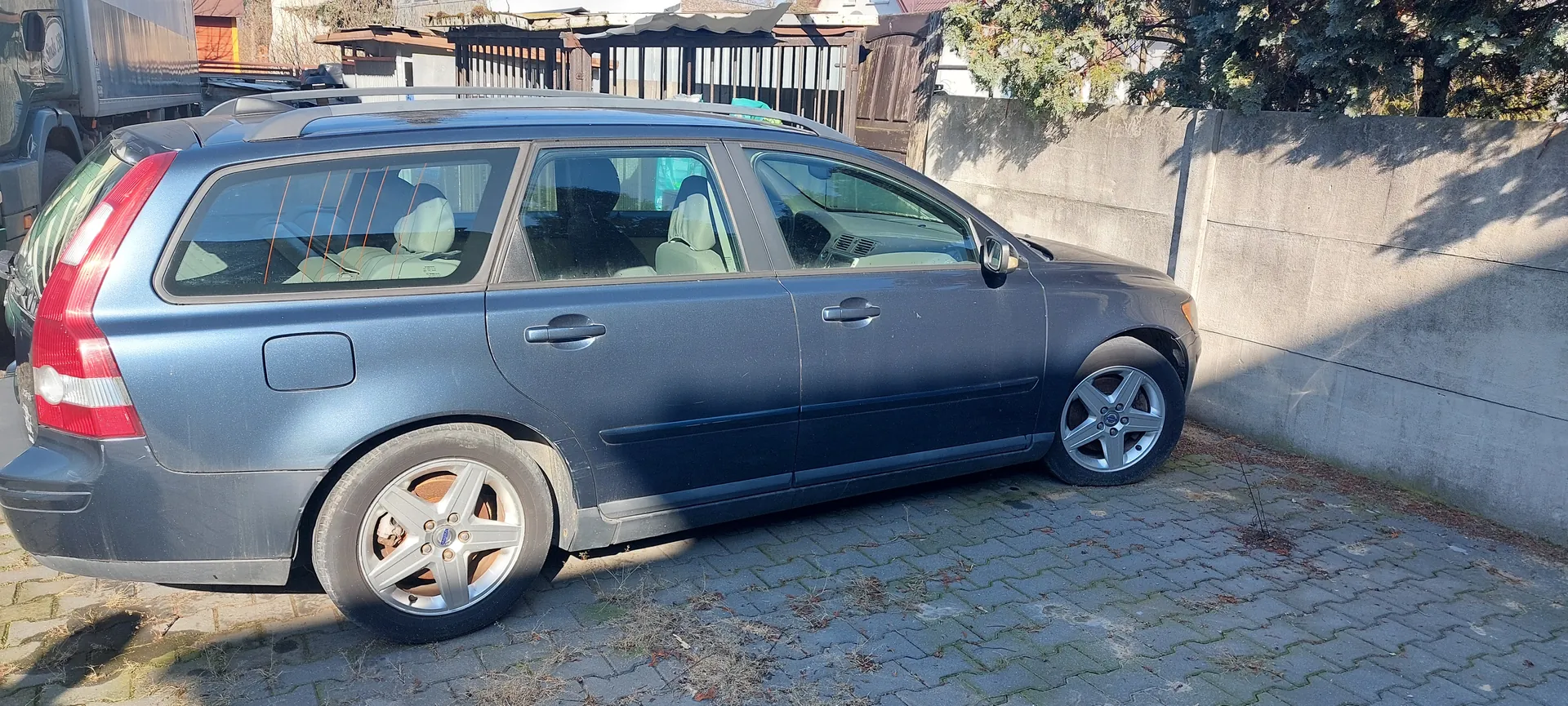 Srebrny samochód typu kombi zaparkowany na brukowanej powierzchni obok betonowej ściany. Po lewej stronie widoczna część innego pojazdu. W tle drewniane ogrodzenie, budynki i drzewa. Samochód ma srebrne felgi, przyciemniane szyby i jasne siedzenia. Na ziemi widoczne są suche trawy i cienie od słońca.