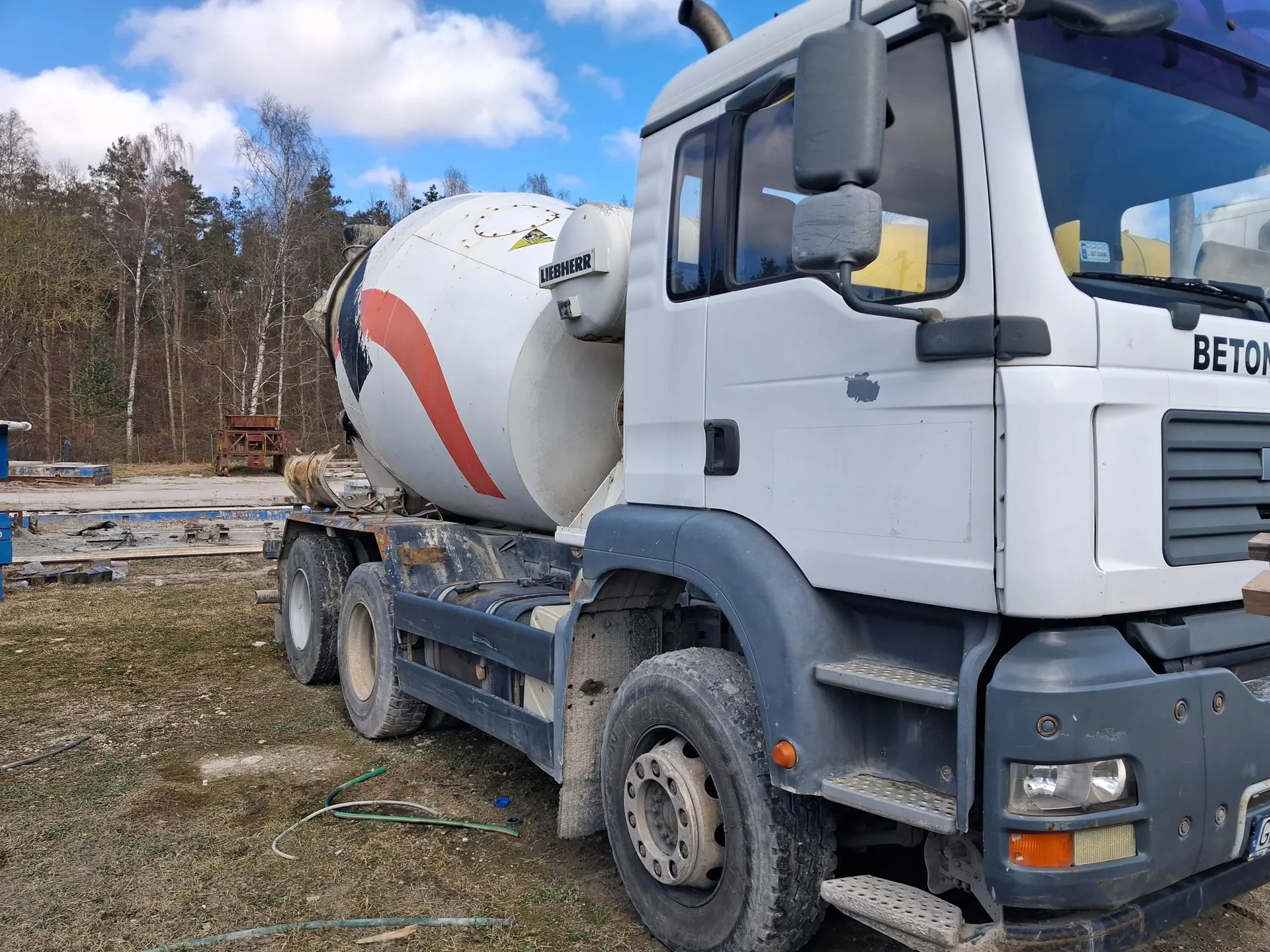 Biały ciężarówka betoniarka marki Liebherr z niebieskim przodem, stojąca na terenie budowlanym. Na boku zbiornika widoczna czerwona linia oraz napis "LIEBHERR", na masce napis "BETON". W tle drzewa, niebo z chmurami, fragmenty materiałów budowlanych i sucha trawa. Na ziemi leżą zielone i białe przewody.