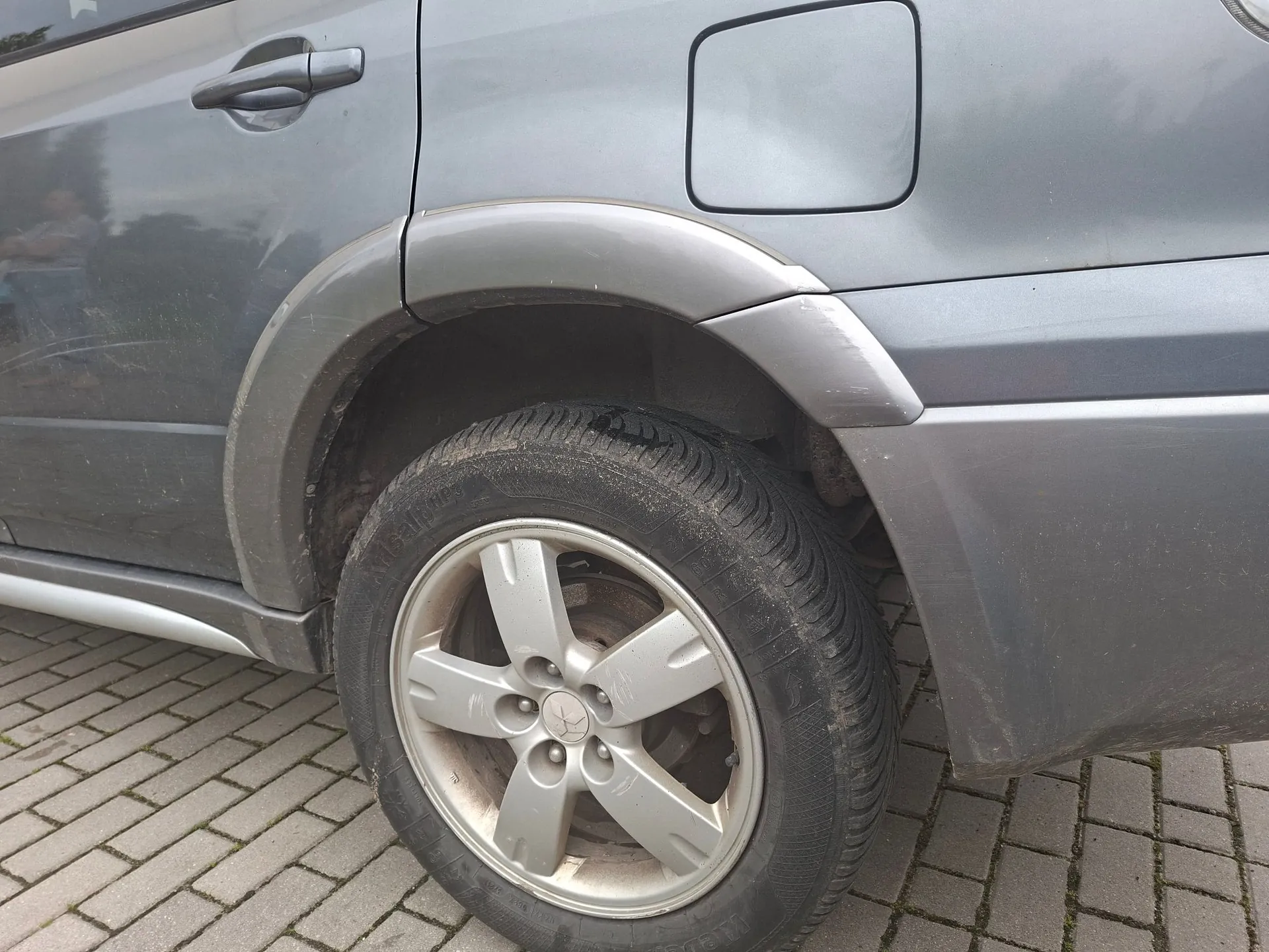 Zbliżenie na tylne lewe koło szarego samochodu zaparkowanego na kostce brukowej. Widoczna jest srebrna felga aluminiowa z pięcioma ramionami oraz opona. Nad kołem znajduje się szary nadkole oraz klapka wlewu paliwa na boku nadwozia.