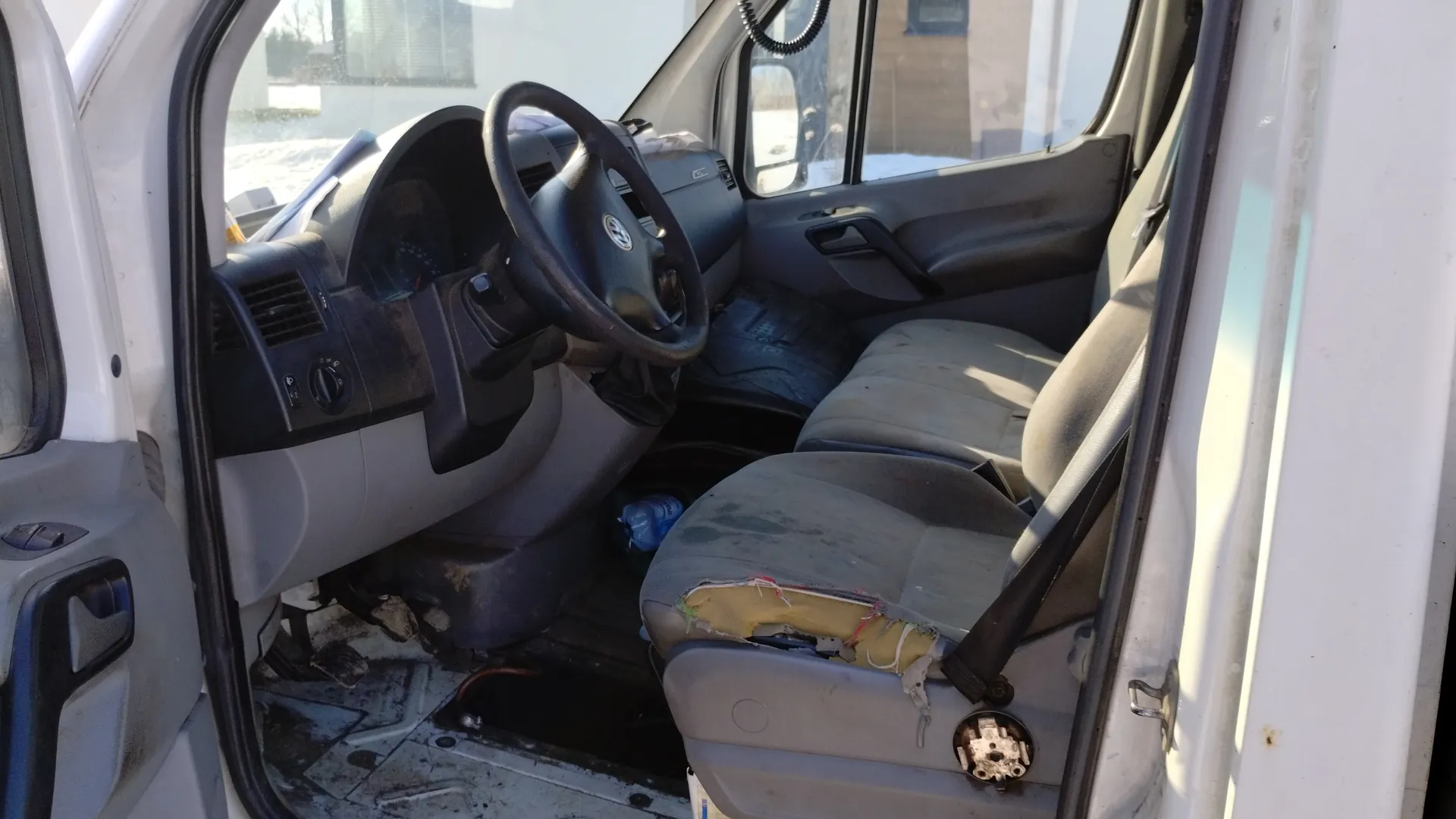 Wnętrze samochodu dostawczego z otwartymi drzwiami. Widoczna kierownica z logo Volkswagen, zniszczone siedzenia (przednie siedzenie pasażera ma rozerwany pokrowiec z odsłoniętą pianką), brudna deska rozdzielcza z przyciskami i wentylacją, oraz zanieczyszczony podłoga. Poza samochodem widać śnieg i fragment budynku. Wnętrze wydaje się nieczyste i zużyte.