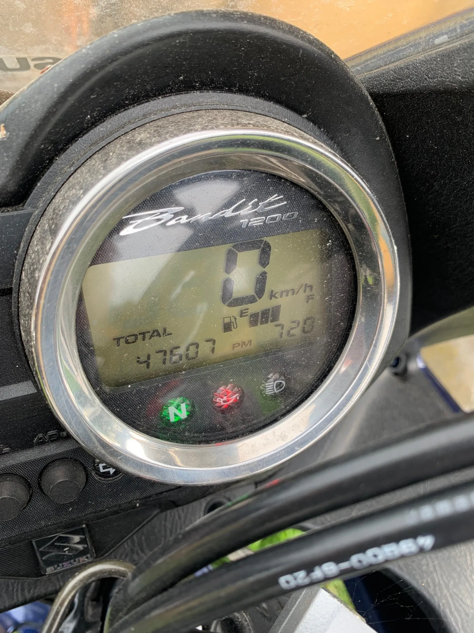 Zbliżenie na cyfrowy licznik motocykla marki Suzuki Bandit 1200. Wyświetlacz LCD pokazuje prędkość 0 km/h, całkowity przebieg 47607 oraz poziom paliwa. Poniżej ekranu znajdują się kontrolki, w tym zielona kontrolka neutralnego biegu z literą 'N'. Całość oprawiona jest w srebrną, okrągłą ramkę.