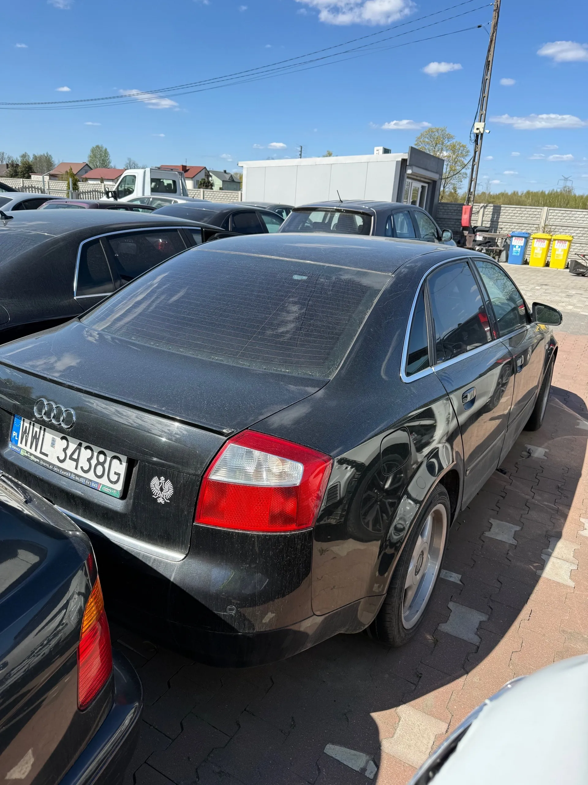 Czarny samochód Audi z tablicą rejestracyjną PL WWL 3438G zaparkowany na placu parkingowym. W tle widoczne są inne pojazdy, budynek, niebieskie i żółte pojemniki na śmieci, słup telekomunikacyjny oraz niebo z chmurami. Na tylnej szybie samochodu znajduje się biały emblemat. Podłoga parkingowa wyłożona jest ceglastymi płytami.