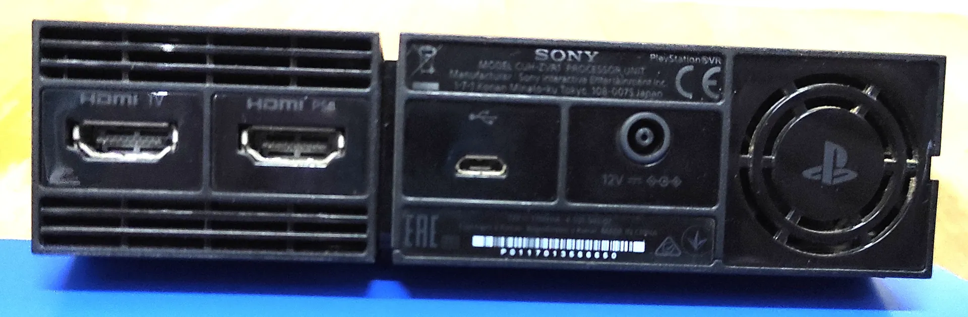 Zdjęcie tylnego panelu konsoli PlayStation VR Processor Unit marki Sony. Widoczne są dwa porty HDMI (oznaczone jako HDMI TV oraz HDMI PS4), port USB, gniazdo zasilania 12V oraz wentylator chłodzący po prawej stronie. Na obudowie znajduje się biały tekst z informacjami technicznymi, w tym model CUH-ZVR1, logo CE oraz oznaczenie EAC.