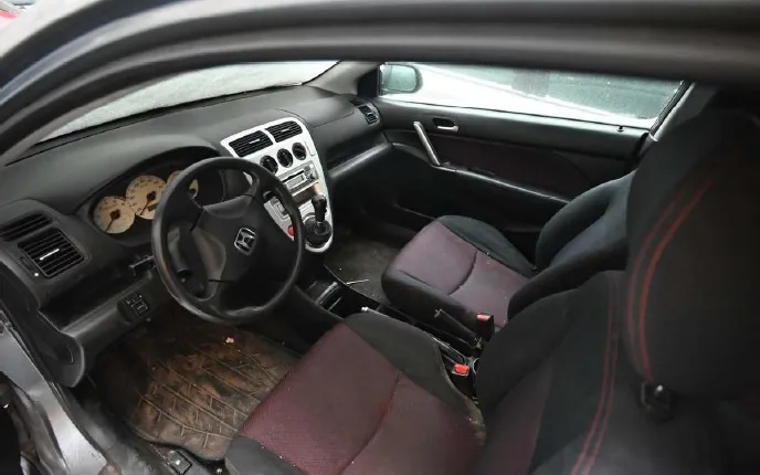 Wnętrze samochodu Honda z widocznym kierownicą z logo marki, tablicą rozdzielczą z analogowymi wskaźnikami, centralną konsolą z systemem audio i klimatyzacją, czarnymi fotelami z czerwonymi przeszyciami oraz zużytymi dywanikami podłogowymi. Widoczne są również drzwi pasażera z uchwytem i szybą, a przez przednią szybę widać zewnętrzne otoczenie z deszczem.