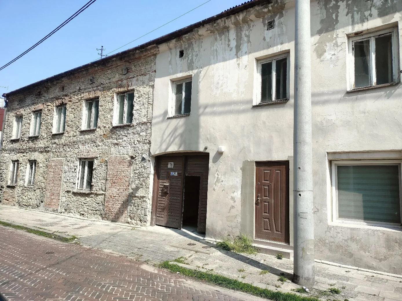 Elewacja starego, dwupiętrowego budynku o dwóch różnych wykończeniach. Lewa część ściany jest z odsłoniętego kamienia i cegły z czterema oknami na górze i trzema na dole. Prawa część budynku jest otynkowana na biało, z dwoma oknami na piętrze, brązowymi drzwiami wejściowymi oraz dużym oknem z roletą na parterze. Obok budynku stoi betonowy słup energetyczny, a przed nim znajduje się chodnik i ulica wyłożona czerwoną kostką brukową.