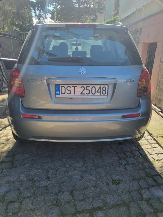 Tylny widok srebrnego samochodu marki Suzuki SX4. Na klapie bagażnika znajduje się logo Suzuki oraz polska tablica rejestracyjna o numerze DST 25048. Samochód stoi zaparkowany na kostce brukowej.