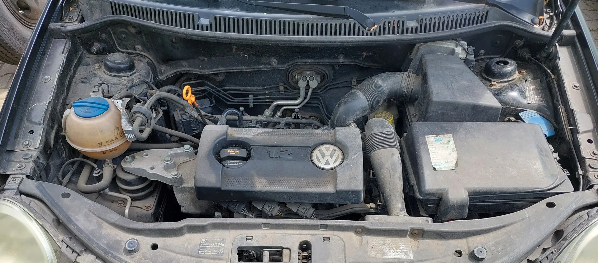 Widok z góry na komorę silnika samochodu marki Volkswagen. Widoczna jest plastikowa osłona silnika z logo VW i oznaczeniem 1.2, zbiornik płynu chłodniczego z niebieską zakrętką po lewej stronie oraz obudowa filtra powietrza po prawej. Elementy silnika są zakurzone.