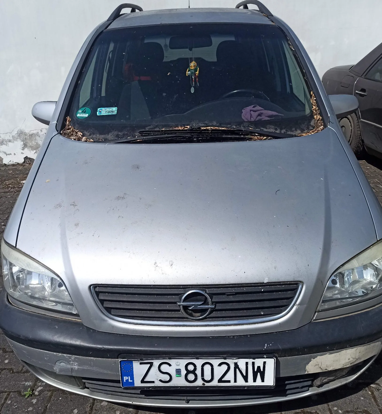 Srebrny samochód marki Opel widziany od przodu. Pojazd ma widoczną tablicę rejestracyjną o treści ZS 802NW. Na przedniej szybie znajdują się naklejki ubezpieczeniowe oraz zawieszka z ozdobą. Na masce i przy wycieraczkach widoczne są zabrudzenia oraz liście.