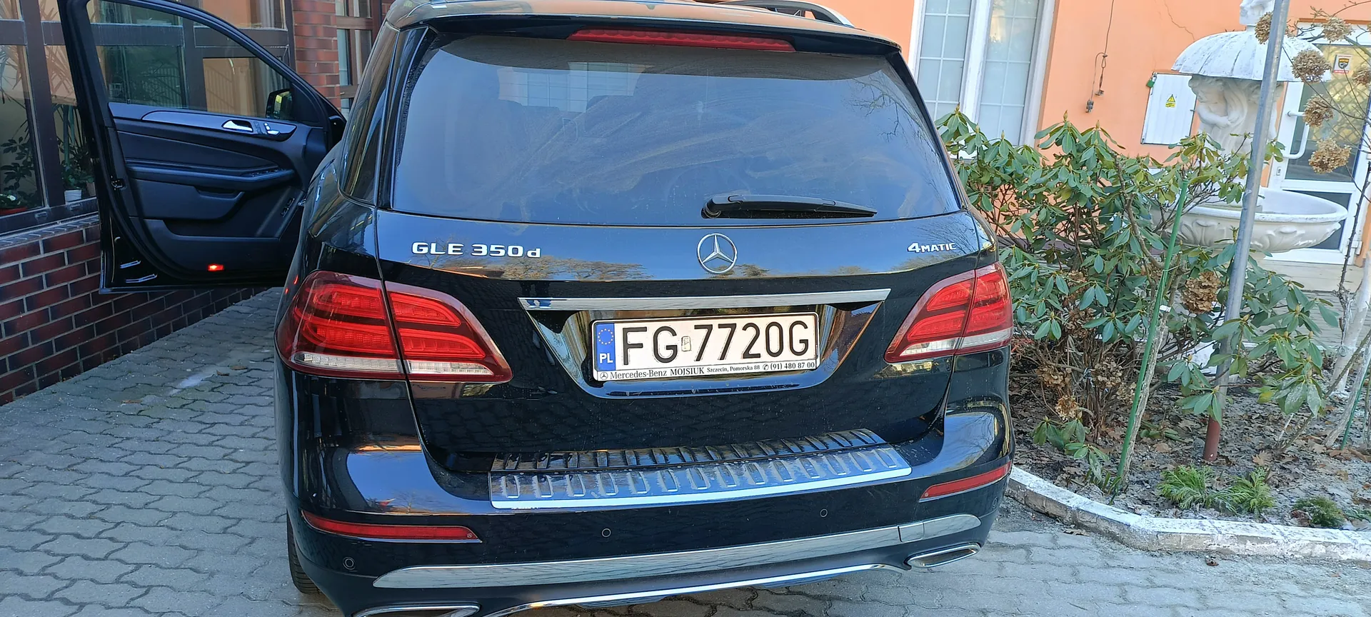 Czarny samochód Mercedes-Benz GLE 350d z widocznym tyłem, tablicą rejestracyjną FG 7720G (PL), napisem 4MATIC po prawej stronie i otwartą lewą przednią drzwiami. Na tablicy widoczny napis: Mercedes-Benz MOJSIUK Szczecin, Pomorska 88, (91) 480 87 00. W tle pomarańczowy budynek z ceglaną ścianą, krzewy, fontanna oraz brukowana powierzchnia.