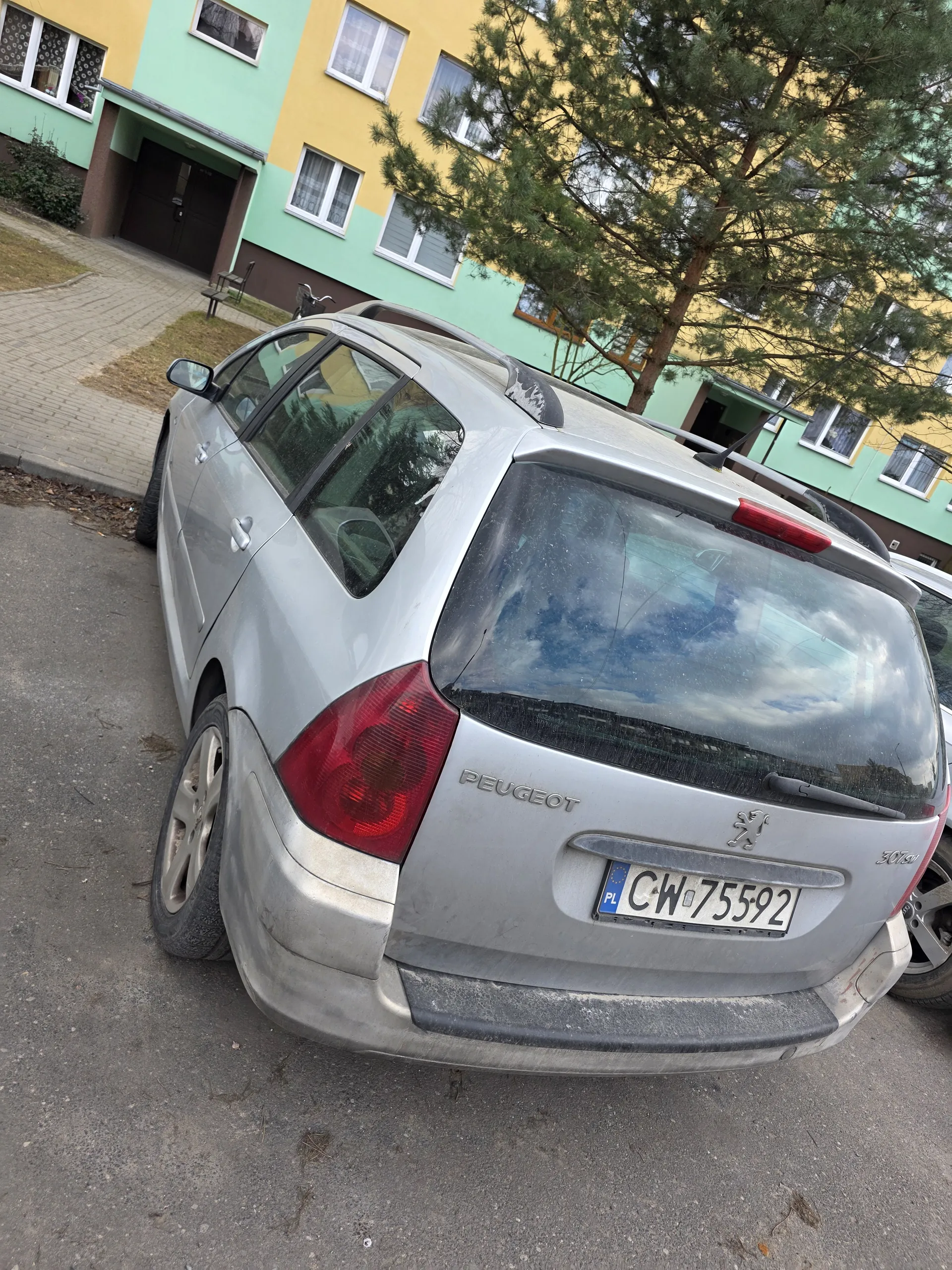 Srebrny samochód marki Peugeot z tablicą rejestracyjną CW75592 zaparkowany na asfaltowej powierzchni. W tle budynki o żółto-zielonych elewacjach z oknami, drzewa i brukowana ścieżka.