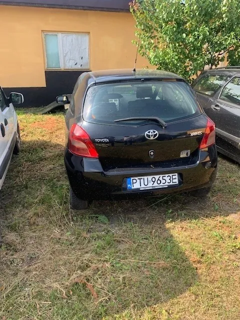 Tylny widok czarnego samochodu marki Toyota Yaris zaparkowanego na trawiastym podłożu. Samochód ma polską tablicę rejestracyjną o numerze PTU 9653E. W tle widoczny jest żółty budynek z oknem oraz zielone drzewo po prawej stronie. Po lewej i prawej stronie widoczne są fragmenty innych pojazdów.