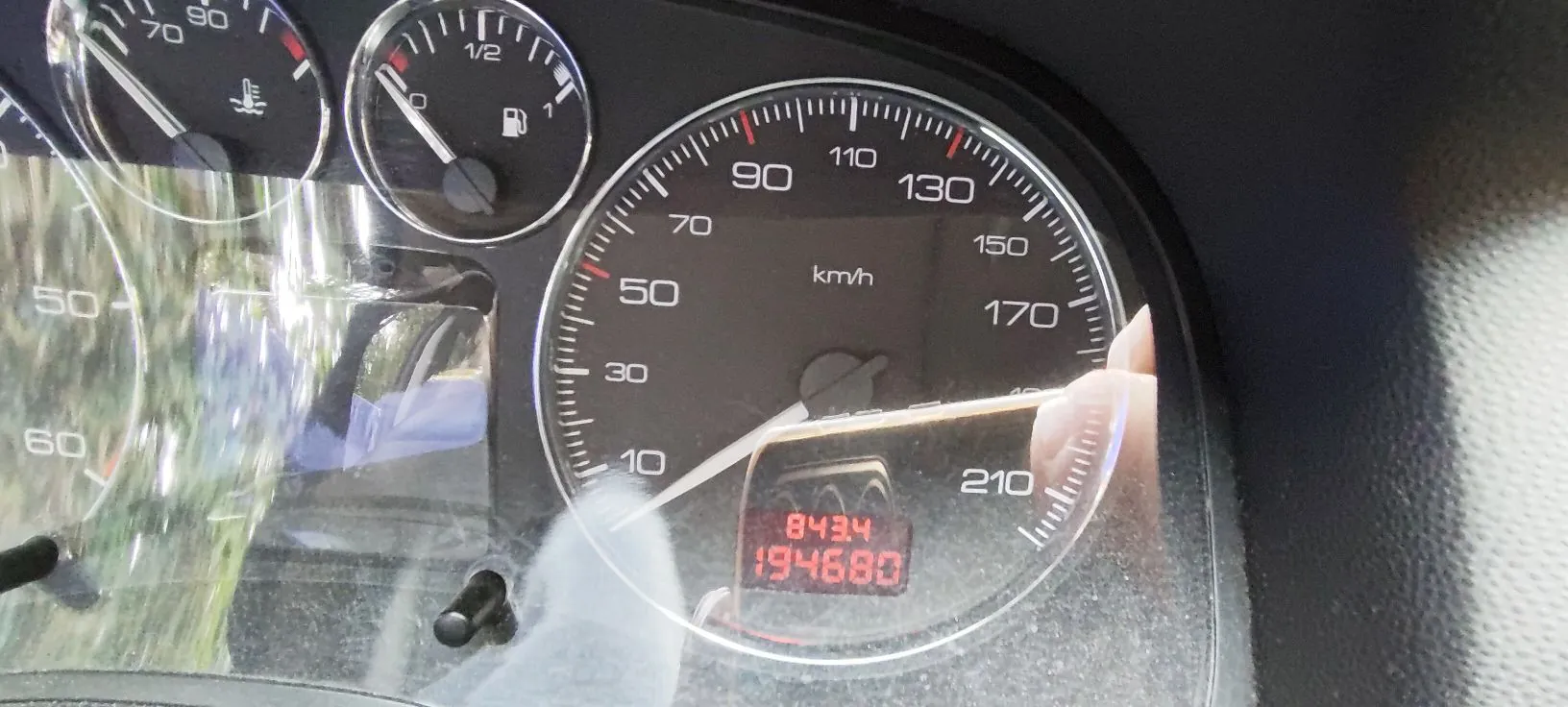 Zbliżenie na deskę rozdzielczą samochodu. Widoczny jest prędkościomierz z podziałką do 210 km/h, wskaźnik paliwa oraz fragment innego wskaźnika po lewej stronie. Na prędkościomierzu znajduje się cyfrowy licznik przebiegu wyświetlający liczbę 194680 oraz mniejszą wartość 843.4 powyżej. Na powierzchni szybki widać odblaski otoczenia.
