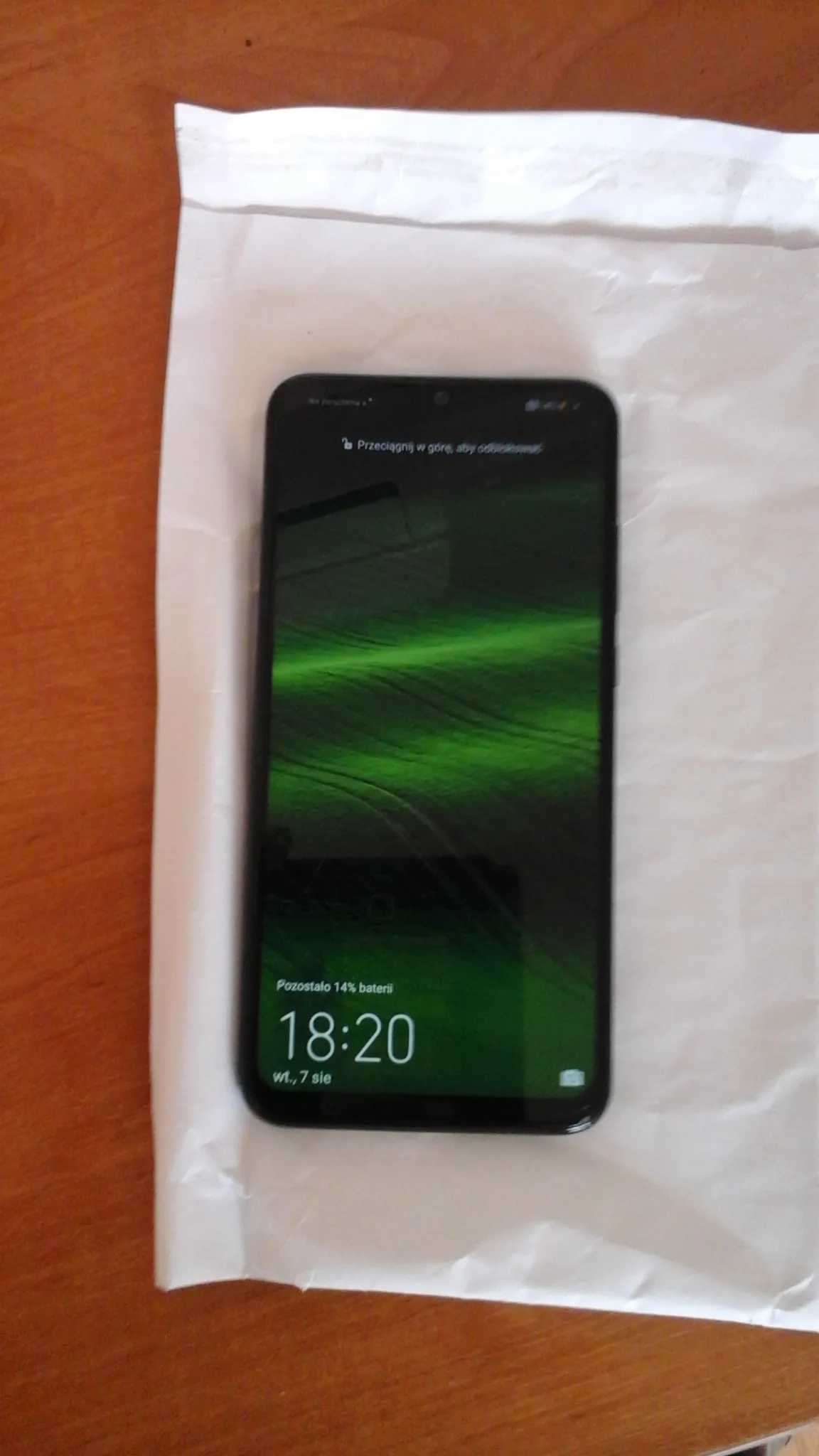 Czarny smartfon leżący poziomo na białym papierze na drewnianym stole. Ekran włączony z zielonym tłem przypominającym zorzę polarną. Na ekranie widoczny czas 18:20, napis "Pozostało 14% baterii", data "Wt, 7 sie" oraz pionowy tekst po lewej stronie: "Przeciągnij w górę, aby odblokować".