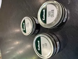 Trzy srebrne słoiki z kremem do butów marki Collonil na ciemnej powierzchni. Etykiety na słoikach zawierają napisy 'Collonil' oraz 'Shoe Cream' w zielonym kolorze na białym tle. Słoiki ułożone są w trójkątnym kształcie.