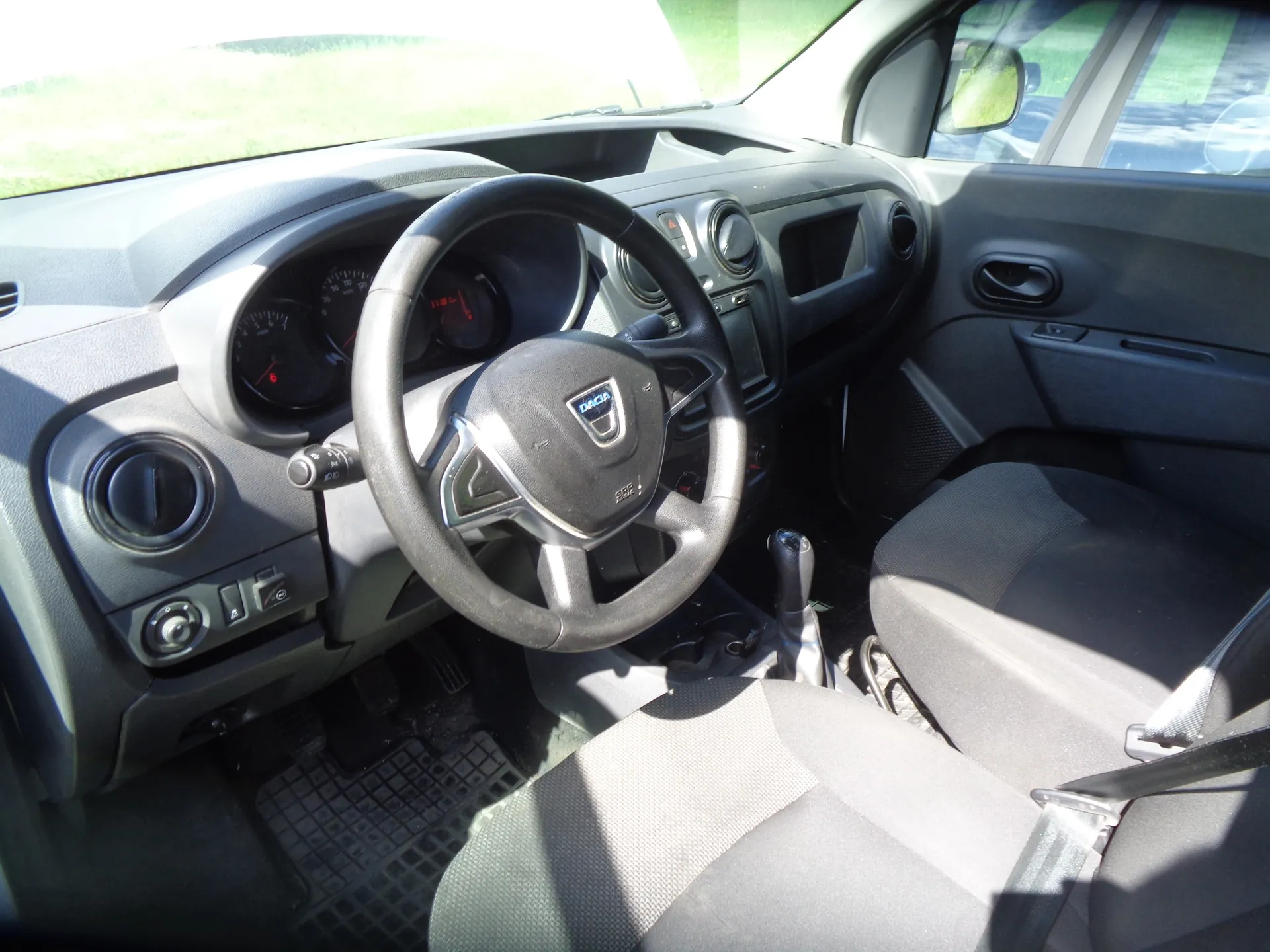 Wnętrze samochodu marki Dacia, widok z perspektywy kierowcy. Widoczna jest czarna kierownica z logo Dacia w centrum, deska rozdzielcza z zegarami, otwory nawiewów oraz ciemnoszare materiałowe fotele. W tle widać fragment przedniej szyby i boczne lustro.