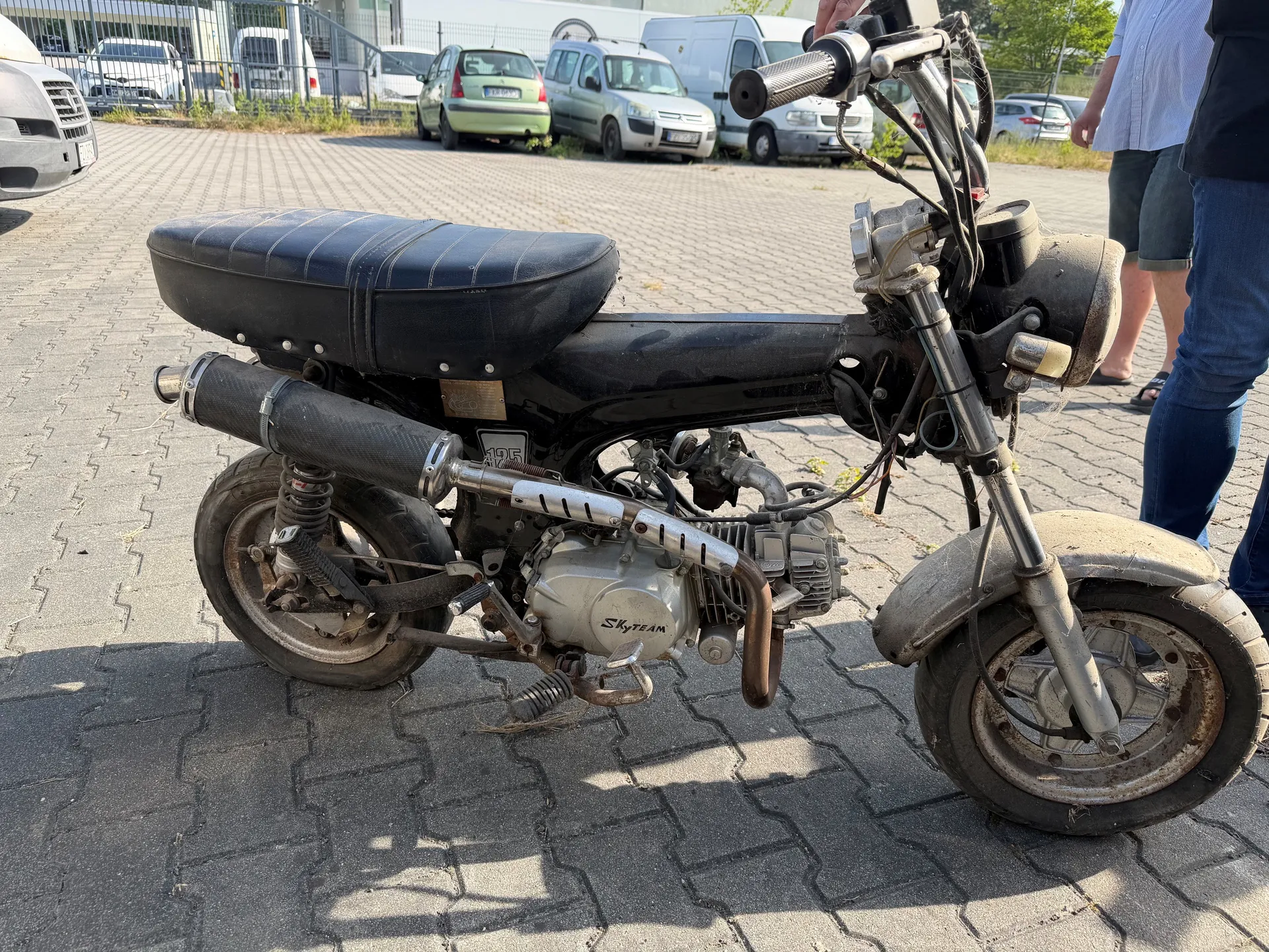 Czarny skuter z silnikiem oznaczonym napisem 'Skyteam' i tablicą rejestracyjną '19F'. Skuter ma czarne siedzenie, wydech oraz zardzewiałe elementy. W tle parking z samochodami (w tym zielonym i srebrnym), ogrodzenie i dwie osoby stojące obok (jedna w niebieskiej koszuli i szortach, druga w dżinsach). Podłoga z kostki brukowej.