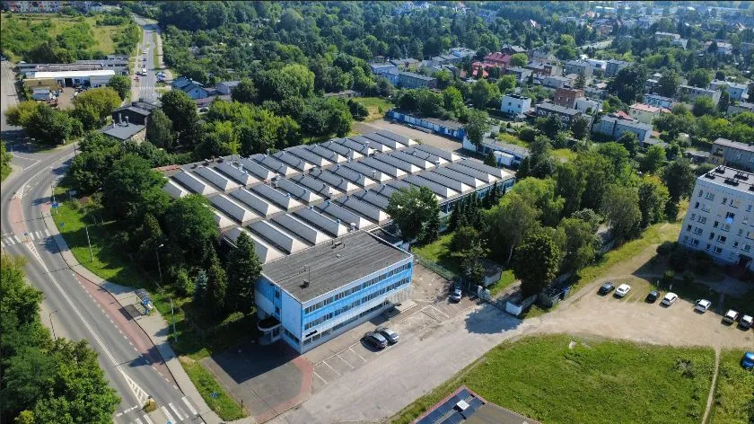 Widok z lotu ptaka na kompleks budynków przemysłowych i mieszkalnych w otoczeniu zieleni. W centrum znajduje się duży budynek z charakterystycznym, wielospadowym dachem o jasnych i ciemnych pasach. Przed nim stoi mniejszy, dwupiętrowy budynek biurowy w kolorze białym i niebieskim z parkingiem przed wejściem. Po lewej stronie przebiega droga z pasami dla pieszych i chodnikiem. W tle widoczne są domy jednorodzinne, drzewa oraz inne zabudowania miejskie.