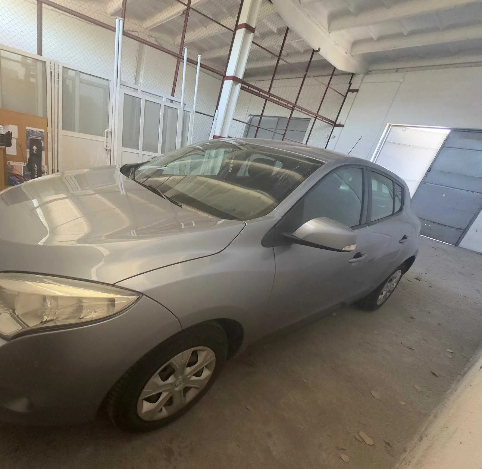 Srebrny samochód osobowy typu hatchback zaparkowany wewnątrz hali. W tle betonowa podłoga, metalowe belki dachu, okna oraz drzwi. Po lewej stronie tablica ogłoszeniowa z papierami.