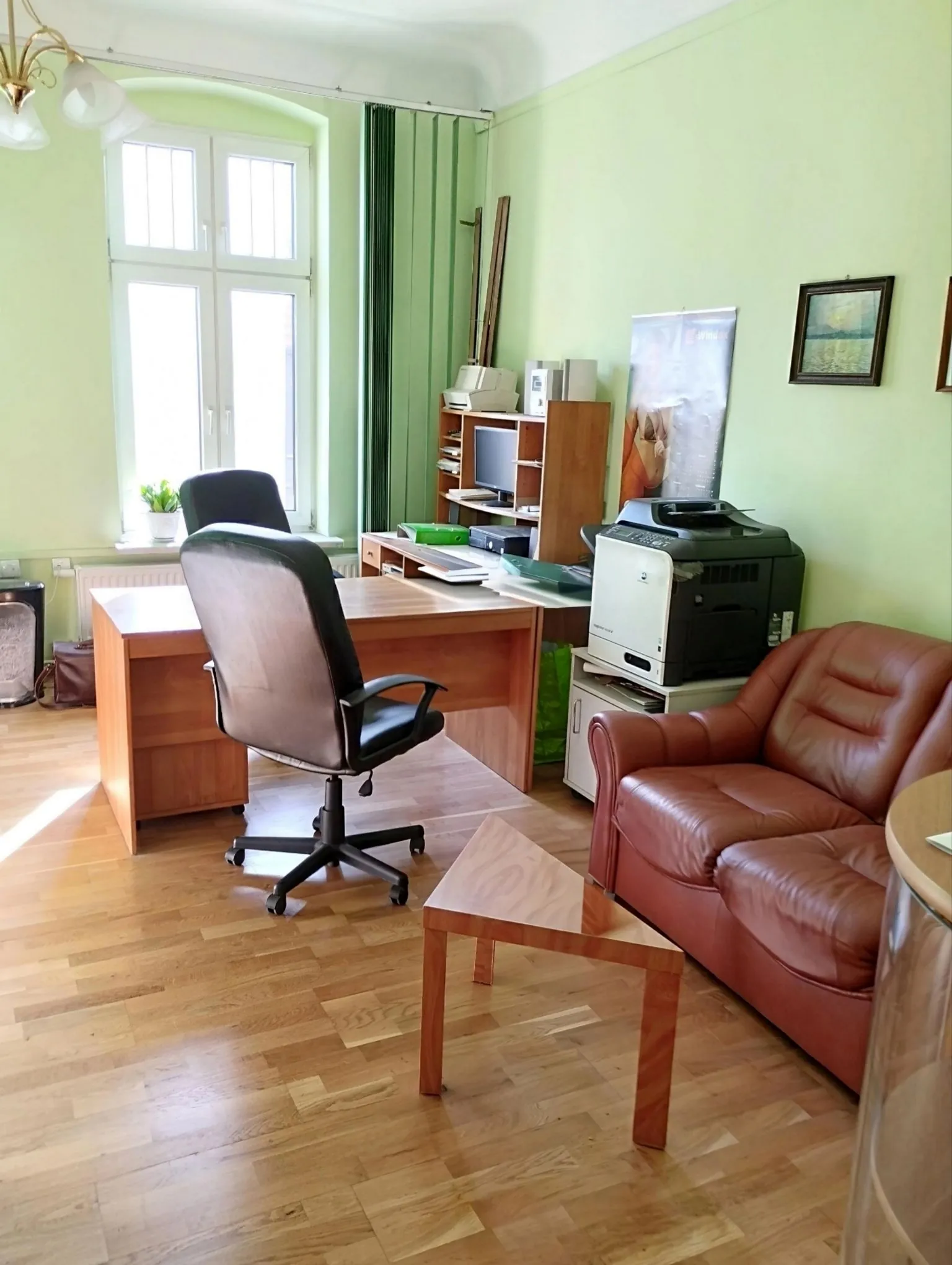 Jasne pomieszczenie biurowe z jasnozielonymi ścianami i drewnianą podłogą. W centrum znajduje się drewniane biurko z czarnym fotelem biurowym. Po prawej stronie stoi brązowa, skórzana kanapa oraz mały drewniany stolik kawowy. W tle widoczny jest regał z monitorem komputera, drukarka wielofunkcyjna oraz okno z białymi ramami. Na ścianie wisi obraz w ciemnej ramie oraz plakat.