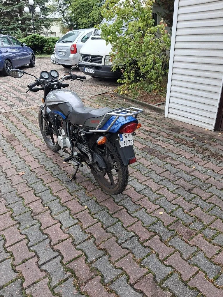 Srebrny motocykl zaparkowany na kostce brukowej. Motocykl jest ustawiony tyłem do widza, widoczna jest tablica rejestracyjna o numerze SY 6770. W tle znajdują się dwa samochody: biały samochód osobowy oraz biały samochód dostawczy, a obok nich zielone krzewy i fragment białej ściany budynku z poziomymi żłobieniami.