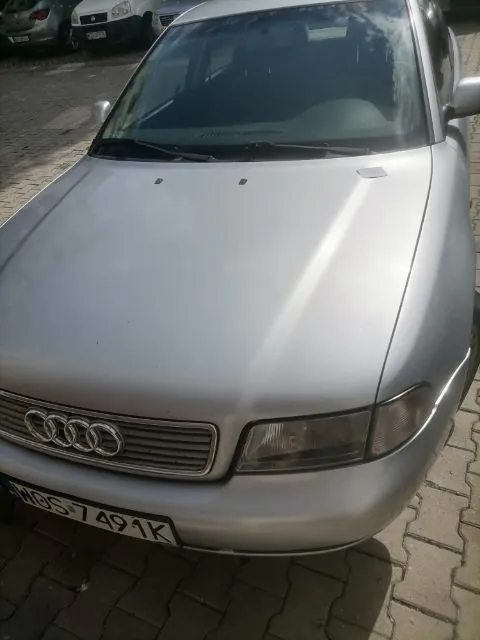 Srebrny samochód marki Audi zaparkowany na kostce brukowej. Widoczny jest przód pojazdu z charakterystycznym grillem z czterema pierścieniami oraz tablicą rejestracyjną o numerze WBS 7491K.