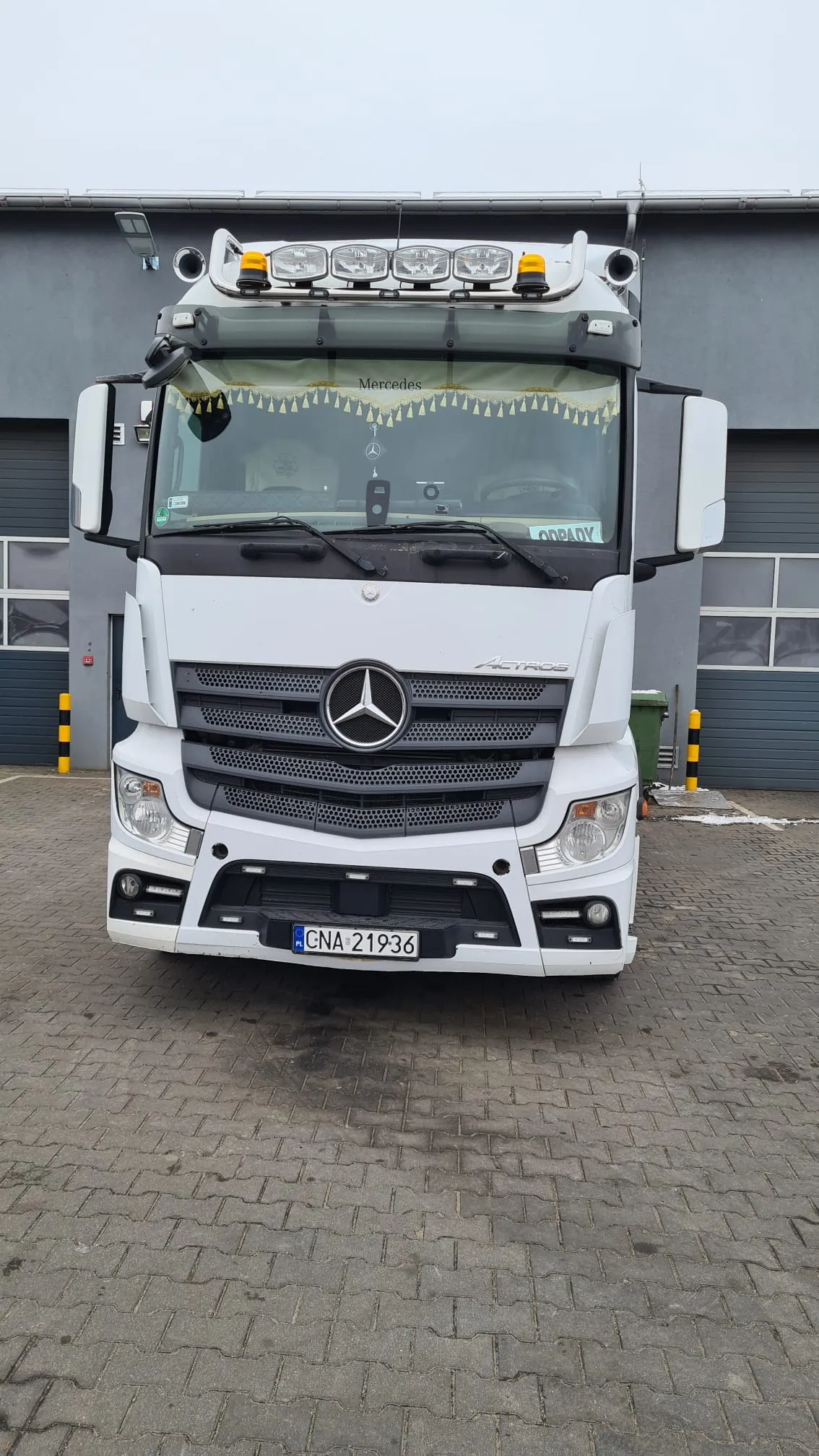 Biały ciągnik siodłowy marki Mercedes-Benz Actros zaparkowany przodem do widza na kostce brukowej przed szarym budynkiem przemysłowym. Pojazd posiada dodatkowe oświetlenie na dachu, tablicę rejestracyjną o numerze GNA 21936 oraz napis 'Mercedes' na górnej części przedniej szyby.