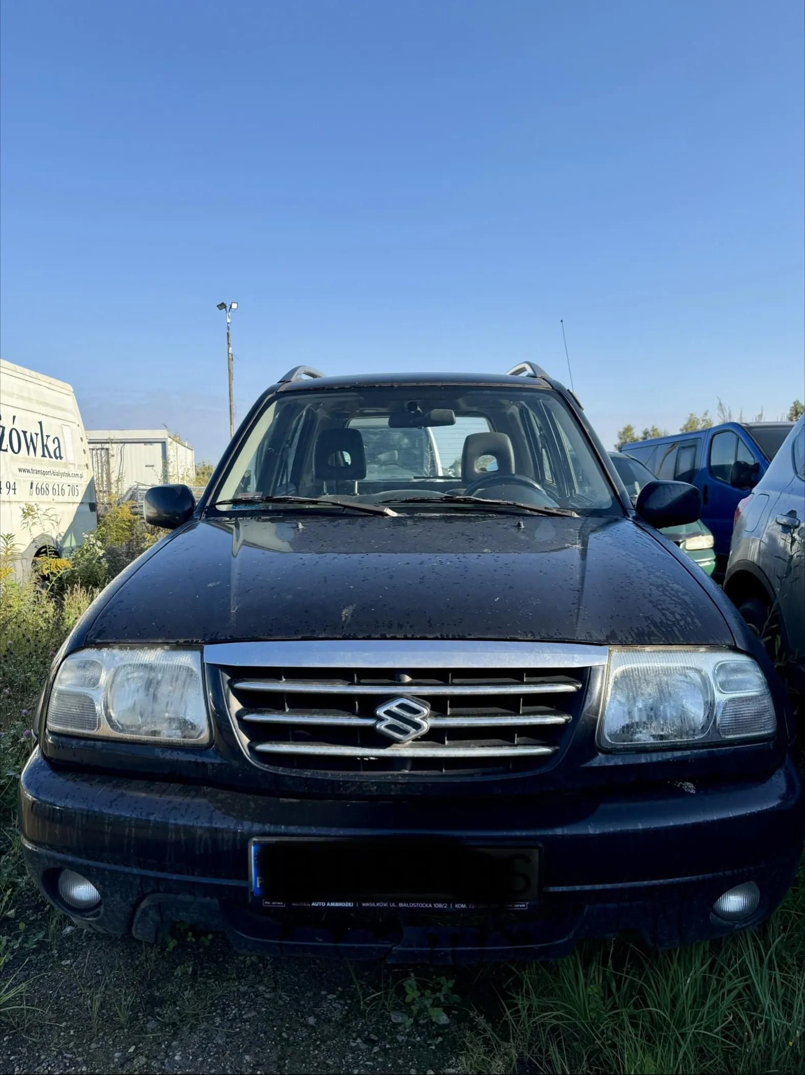 Widok z przodu na czarny samochód marki Suzuki zaparkowany na trawiastym terenie. Pojazd jest zakurzony. W tle widoczna jest biała naczepa z napisem 'żówka' oraz numerem telefonu '94 668 616 703', niebieski samochód dostawczy oraz jasne, bezchmurne niebo.