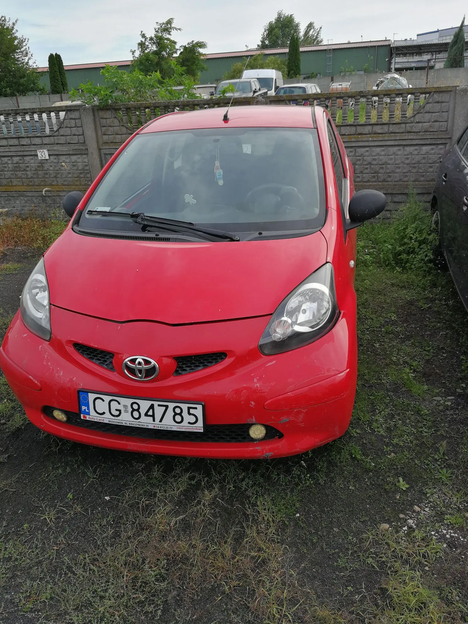 Czerwony samochód marki Toyota zaparkowany na nieutwardzonym terenie. Pojazd posiada polską tablicę rejestracyjną o numerze CG 84785. W tle widoczny jest betonowy płot, zieleń oraz inne zaparkowane samochody.