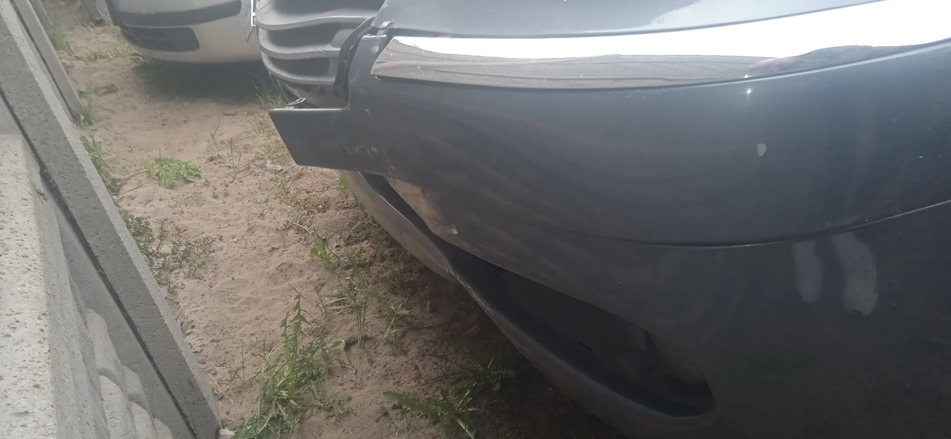 Zbliżenie na uszkodzony przedni zderzak ciemnoszarego samochodu. Plastikowy element zderzaka jest pęknięty i częściowo oderwany, odsłaniając wewnętrzne części pojazdu. Samochód stoi na piaszczystym podłożu z kępkami trawy, obok betonowego ogrodzenia.