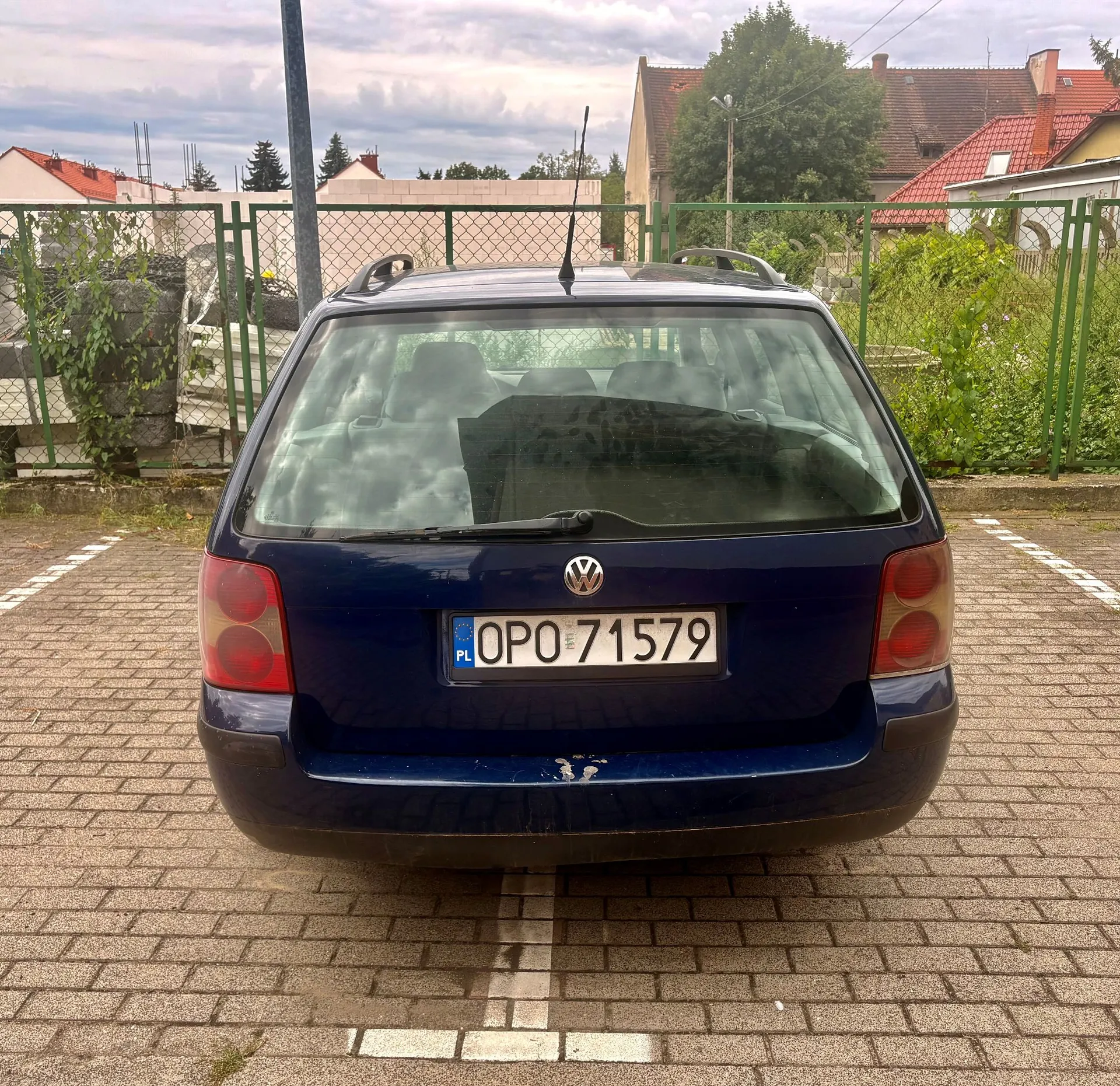 Tylny widok ciemnoniebieskiego samochodu marki Volkswagen w nadwoziu kombi, zaparkowanego na kostce brukowej. Samochód ma polską tablicę rejestracyjną o numerze OPO 71579. W tle widoczny jest zielony płot, roślinność oraz dachy domów pod zachmurzonym niebem.