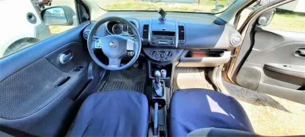 Wnętrze samochodu z widocznym kierownicą z logo Nissan, deska rozdzielcza z radiem, dźwignią zmiany biegów i niebieskimi fotelami. Drzwi po stronie kierowcy są otwarte, a na zewnątrz widoczny jest fragment innego pojazdu i trawa. Wnętrze posiada ciemnoszare panele i dywaniki na podłodze.