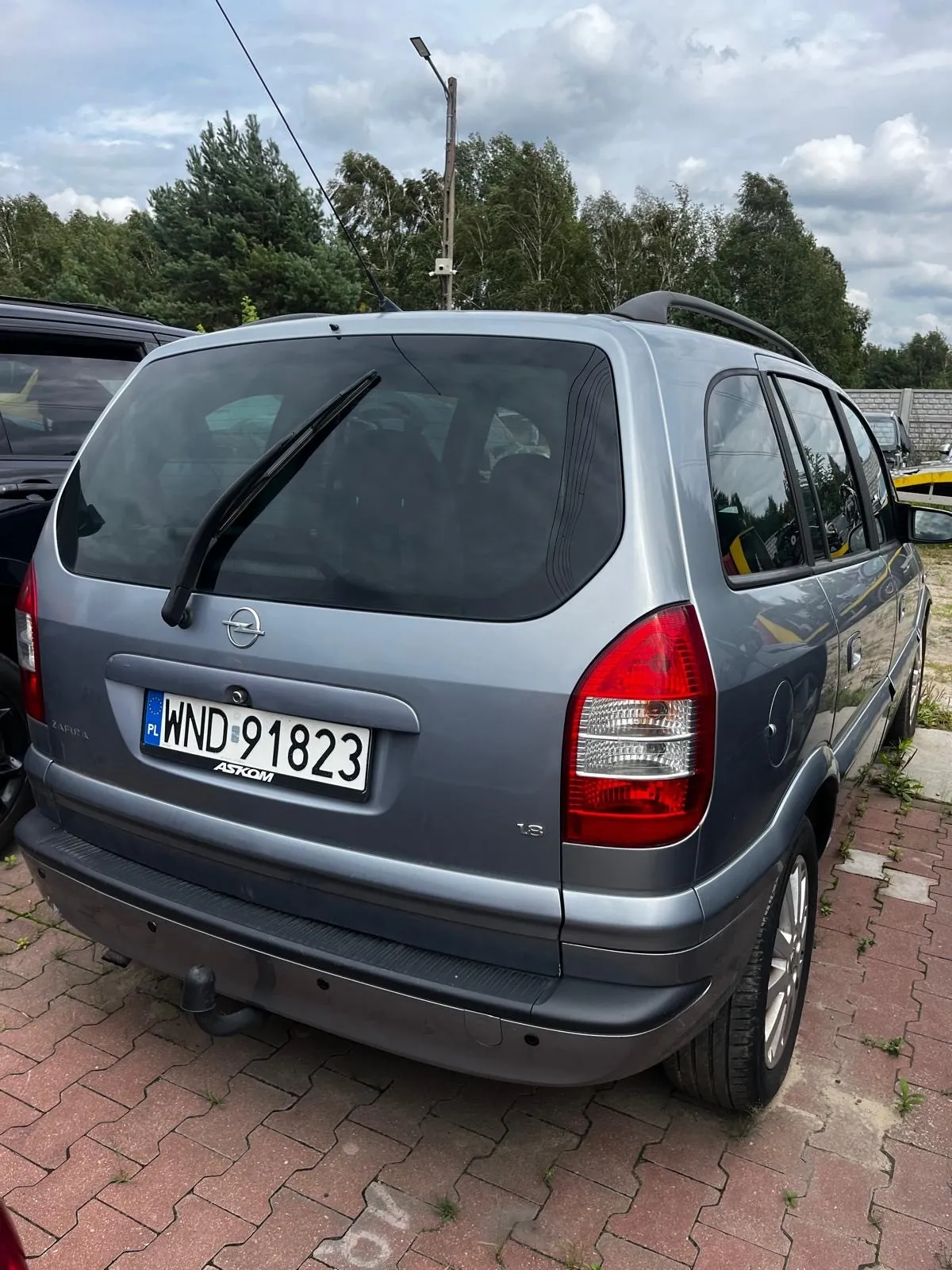Tylny widok srebrnego samochodu marki Opel Zafira zaparkowanego na kostce brukowej. Na klapie bagażnika widoczna jest polska tablica rejestracyjna o numerze WND 91823 oraz napis ASKOM na ramce tablicy. Po lewej stronie znajduje się napis ZAFIRA, a po prawej oznaczenie wersji wyposażenia LS. W tle widoczne są zielone drzewa i zachmurzone niebo.