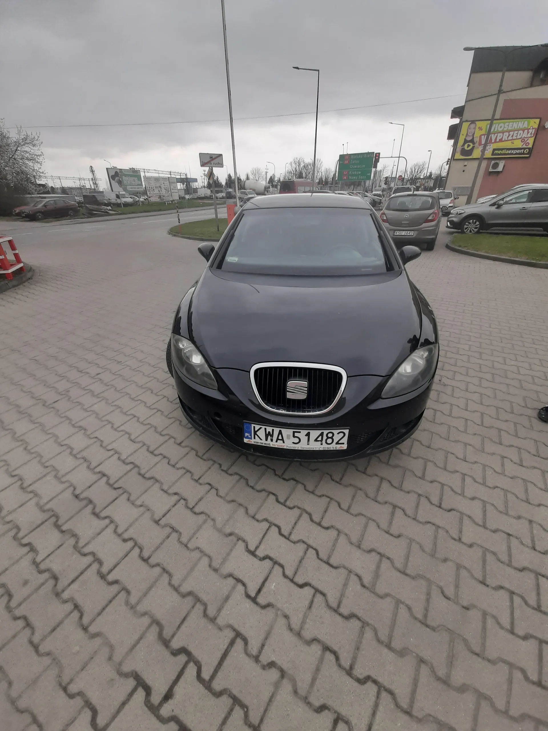Czarny samochód marki SEAT z tablicą rejestracyjną KWA 51482 zaparkowany na brukowanej powierzchni. W tle inne pojazdy, budynki, tablice reklamowe z napisem WYPRZEDAŻ oraz znaki drogowe. Niebo jest zachmurzone.