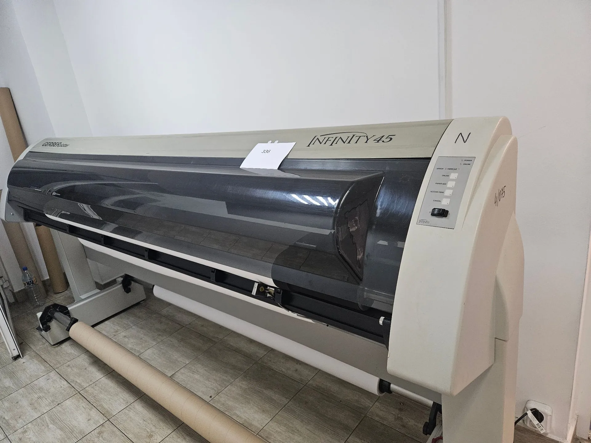 Szeroki ploter do druku wielkoformatowego marki Infinity 45 w kolorze białym i szarym. Urządzenie posiada ciemnoszarą, półprzezroczystą osłonę górną. Po prawej stronie znajduje się panel sterowania z przyciskami i napisem 'Infinity'. Na górnej obudowie przyklejona jest mała biała etykieta z numerem '338'. Pod ploterem widoczna jest rolka z beżowym papierem.