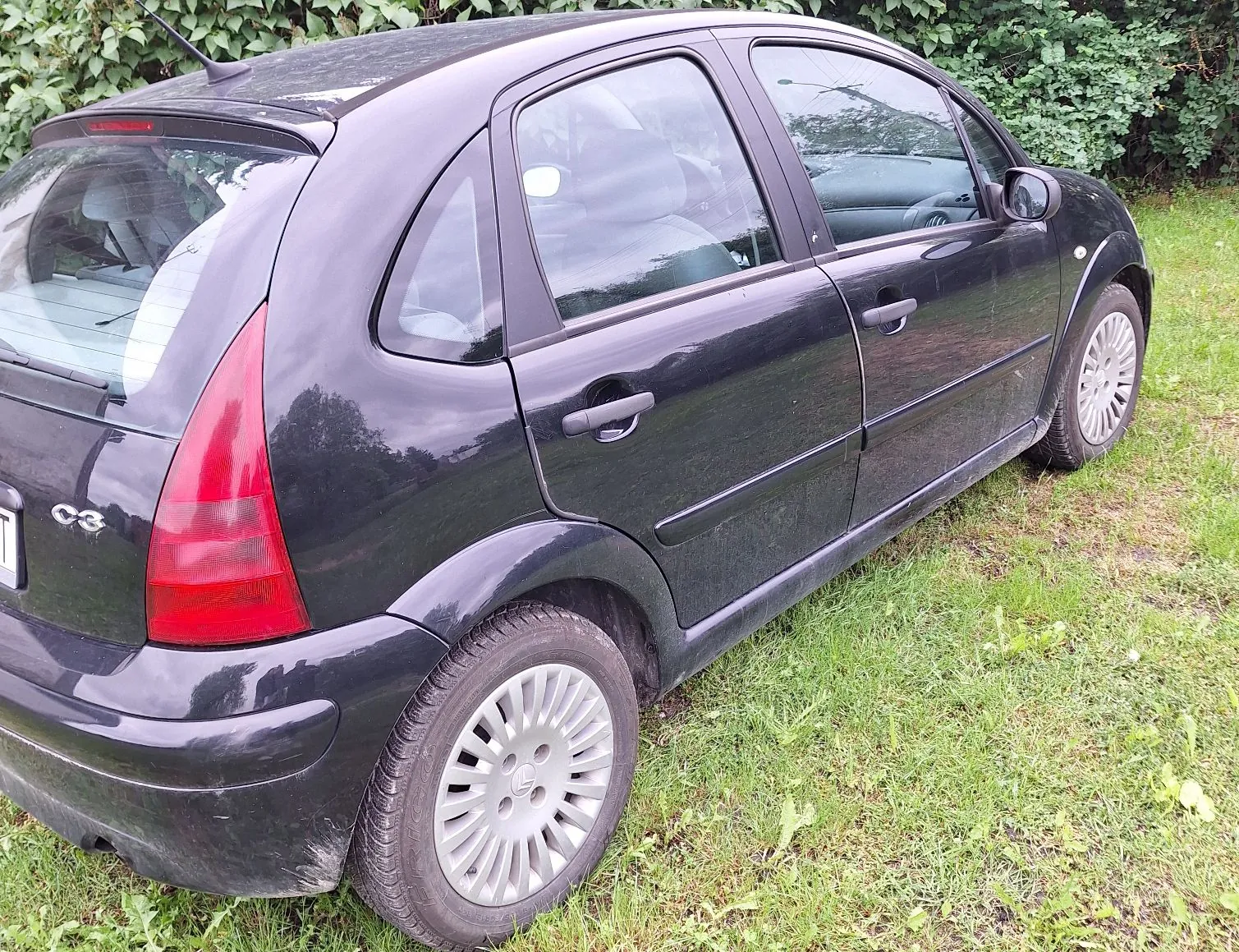 Czarny samochód osobowy marki Citroën C3 zaparkowany na trawie. Widok z boku i z tyłu pojazdu. Na klapie bagażnika widoczny jest napis 'C3'. W tle znajdują się zielone krzewy i drzewa.