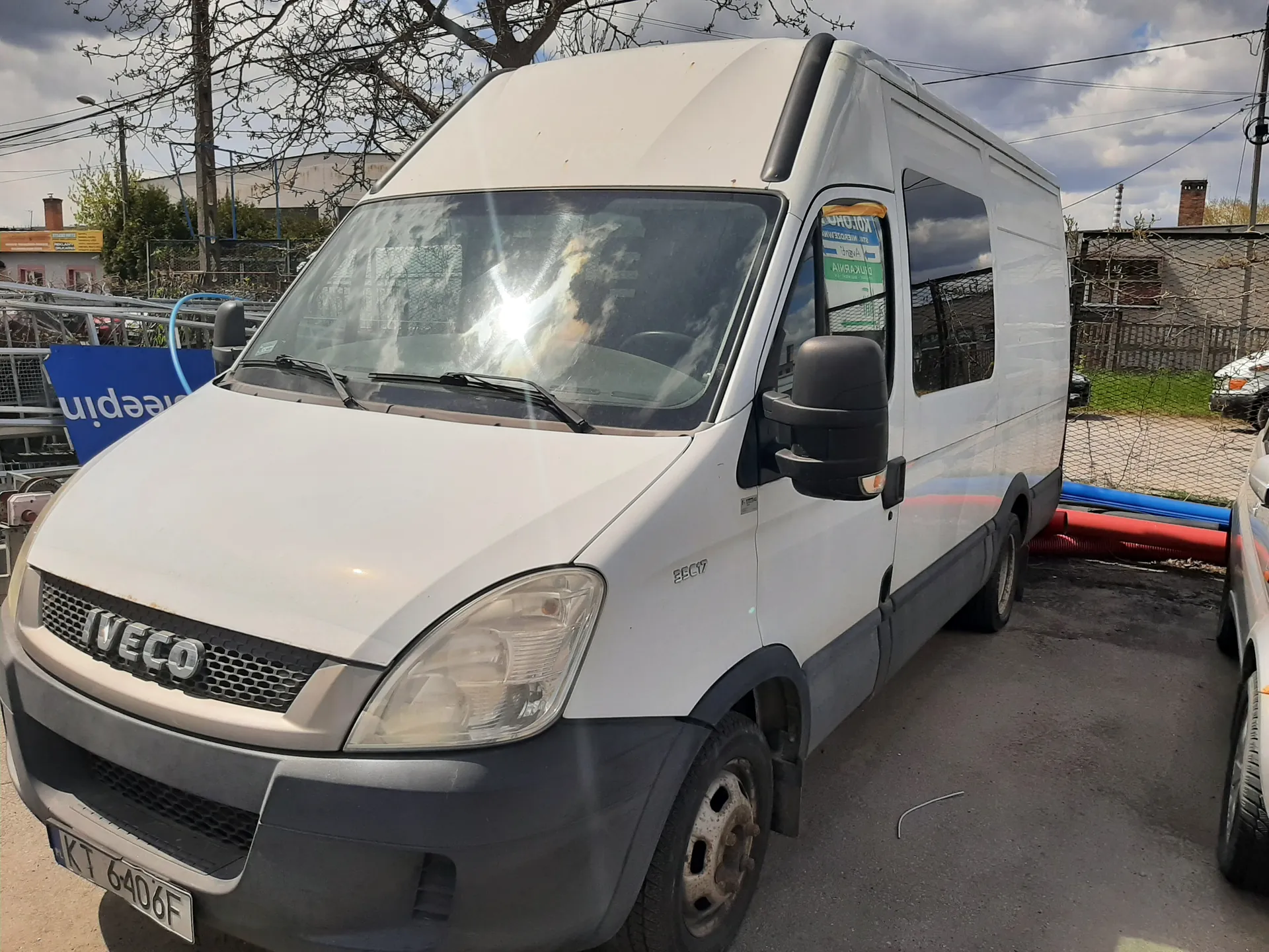 Biały samochód dostawczy marki IVECO zaparkowany na asfaltowej powierzchni. Widoczna tablica rejestracyjna KT 6406F. W tle budynki, ogrodzenie siatkowe, drzewa bez liści oraz niebo z chmurami. Po lewej stronie niebieski znak z częściowo widocznym napisem 'keepin'. Na przedniej szybie odbicie słońca. Po prawej stronie fragment innego pojazdu.