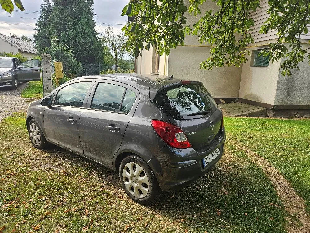 Szary samochód osobowy marki Opel Corsa zaparkowany na trawniku obok jasnego budynku. Widoczny jest tył i bok auta z tablicą rejestracyjną o numerze SCI 39455. W tle widać inny samochód, drzewa oraz fragment domu.