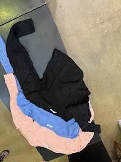 Trzy swetry w kolorach różowym, niebieskim i czarnym ułożone na ciemnej powierzchni. W tle widoczna betonowa podłoga.