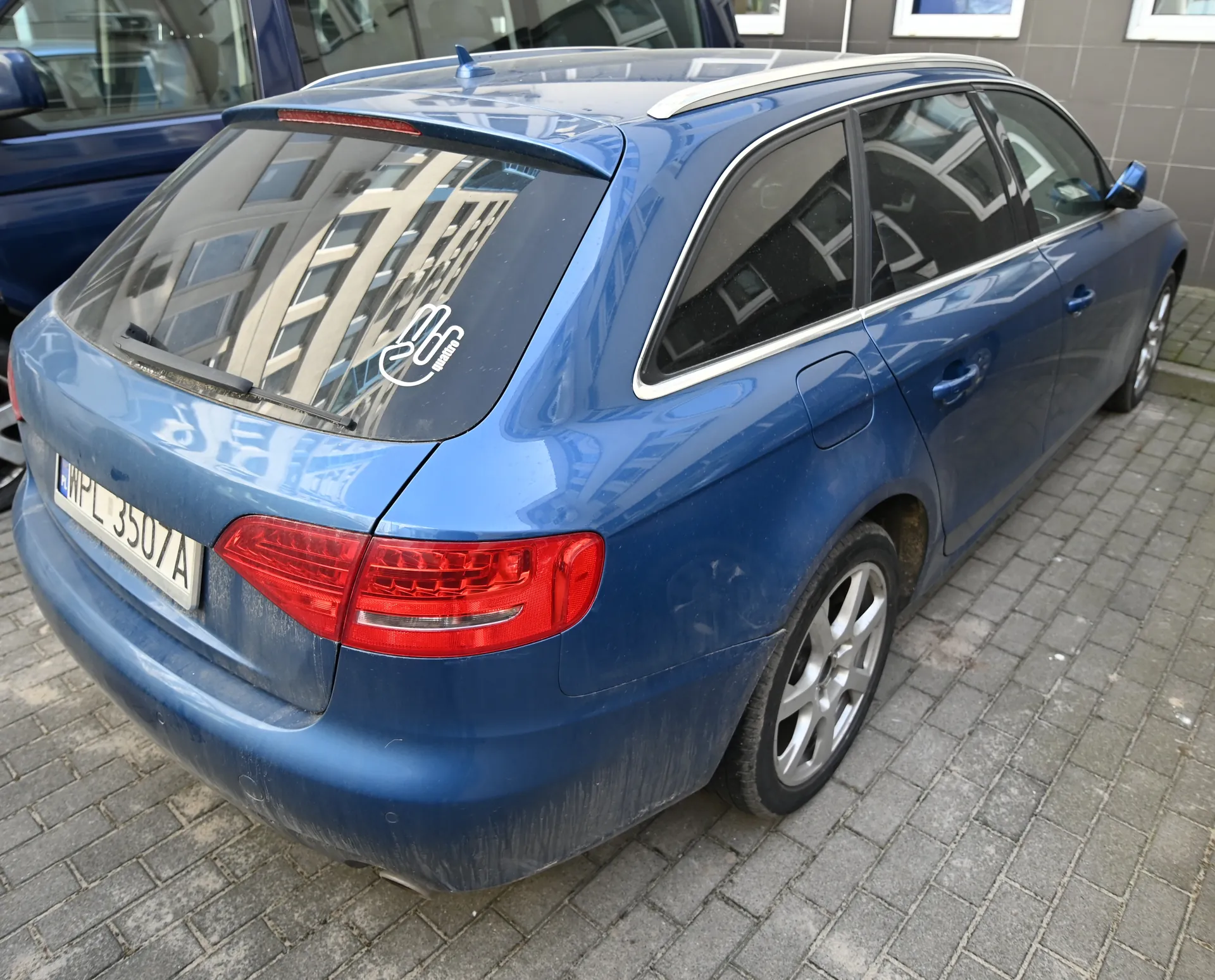 Niebieski samochód typu kombi zaparkowany na brukowanej powierzchni. Widok z tyłu i boku. Tablica rejestracyjna z napisem WPL 3507A. Na tylnej szybie naklejka z białym napisem 200 i dodatkowym tekstem. Samochód posiada dachówkę i przyciemniane szyby. W tle fragment budynku z oknami oraz częściowo widoczny inny niebieski samochód.