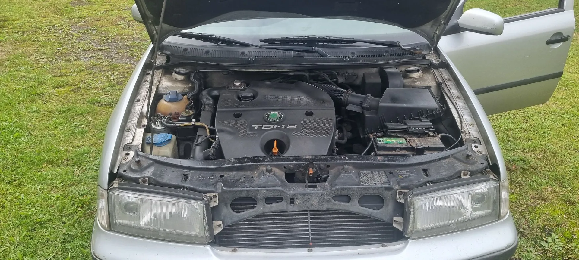 Widok z góry na otwartą maskę srebrnego samochodu, ukazującą komorę silnika. Na czarnej osłonie silnika widnieje napis 'TDI-1.9' oraz logo marki Skoda. Po lewej stronie widoczne są zbiorniki z płynami eksploatacyjnymi, a po prawej skrzynka bezpieczników i obudowa filtra powietrza. Samochód stoi na trawiastym podłożu, a drzwi pasażera są otwarte.