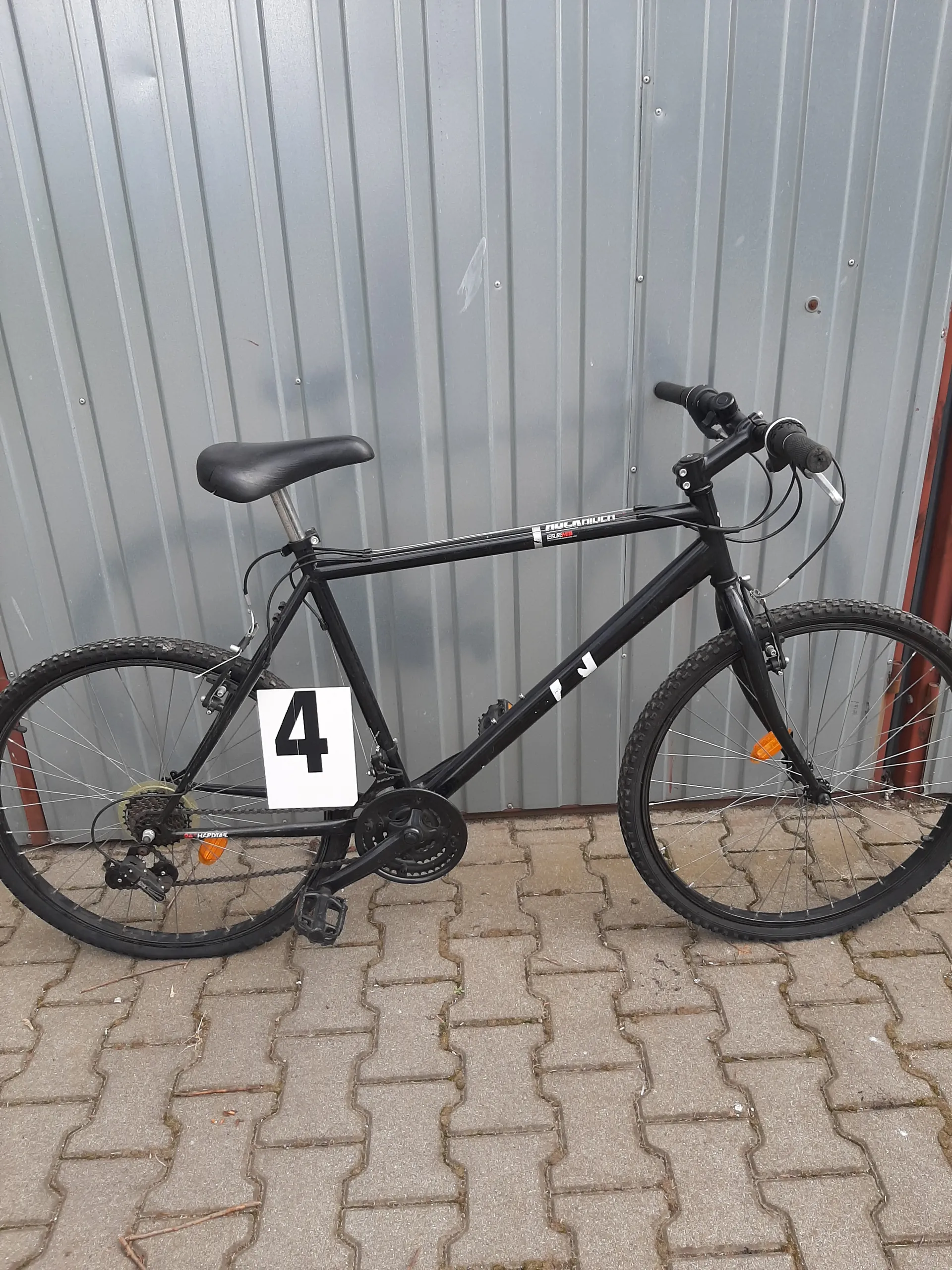 Czarny rower w poziomej orientacji oparty o szarą metalową ścianę z pionowymi listwami i brukowaną powierzchnię z kamiennymi płytami. Na ramie roweru widoczny biały znak z czarną cyfrą 4. Rower ma czarne koła, siedzisko i kierownicę z czarnymi uchwytami.
